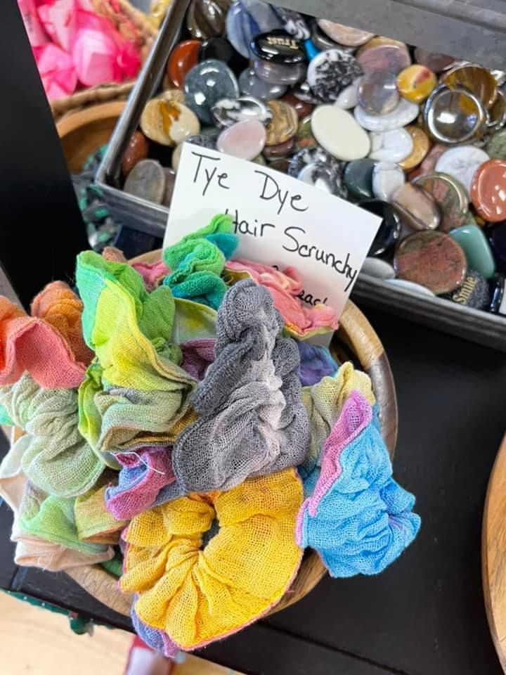 Bowl of tie-dye hair scrunchies with a sign. Background of various colorful stones in a tray.