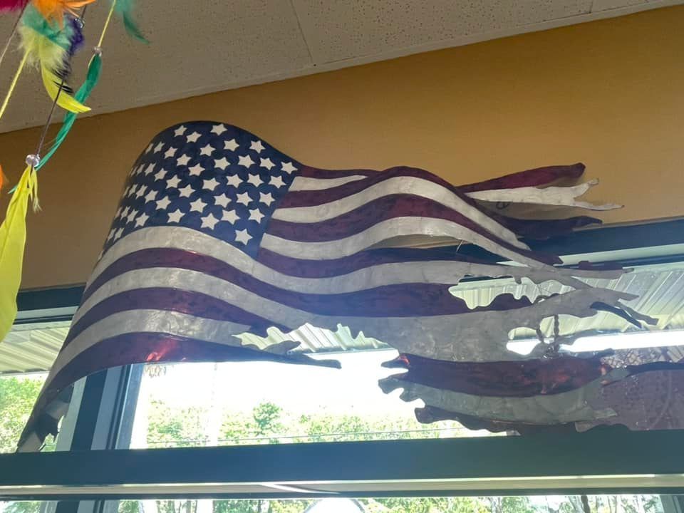 American flag sculpture, torn and waving above a window, mounted on a beige wall.
