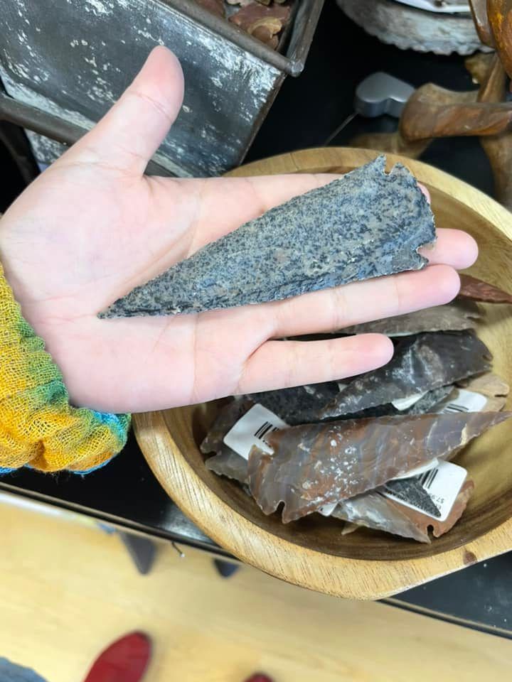 Hand holding a gray, triangular stone above a wooden bowl with other similar stones; a store setting.
