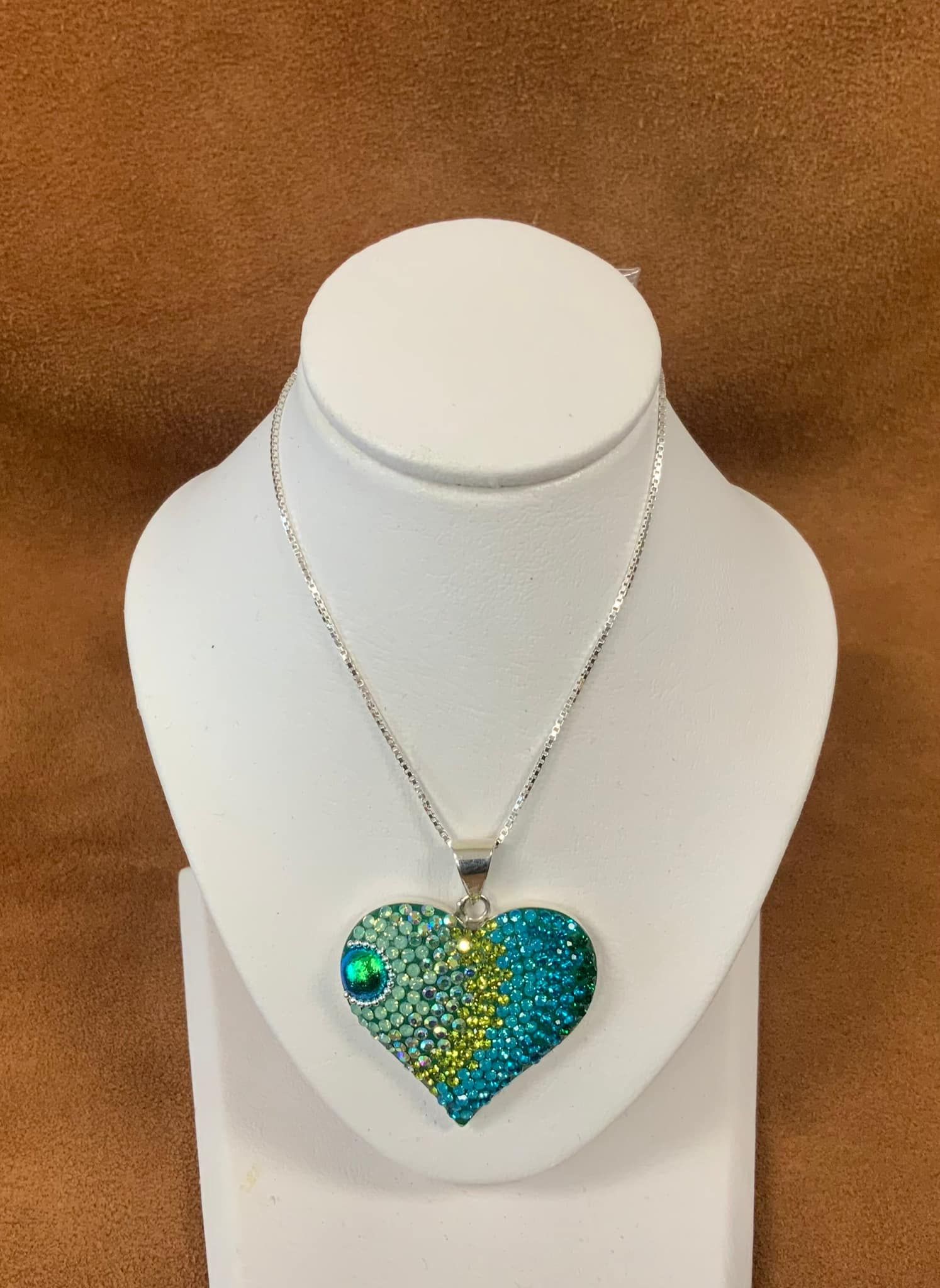 Heart-shaped pendant necklace with blue and green gems on a white display stand, against a brown background.