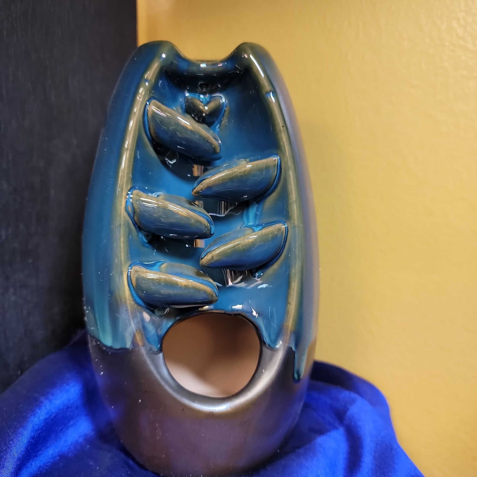 Teal and brown ceramic incense burner with cascading leaf design. Blue cloth background, yellow wall.