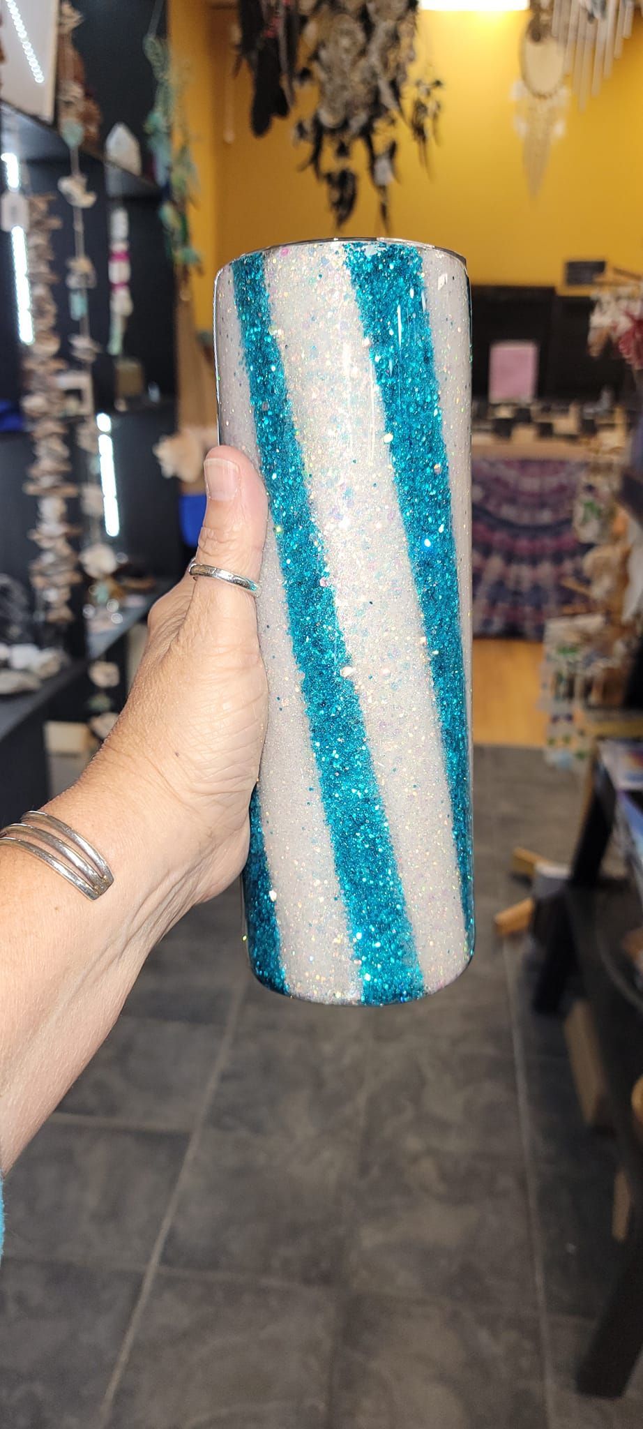 A hand holding a cylindrical glitter tumbler with diagonal blue and white stripes. In a shop.