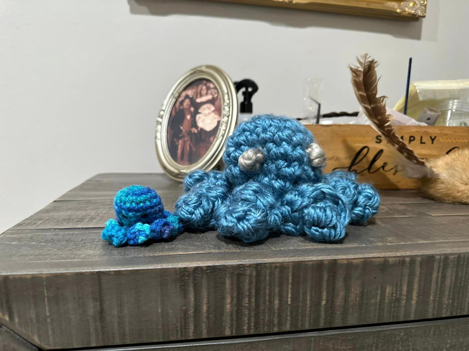 Two blue crocheted octopuses sit on a dark wooden surface, with a framed photo in the background.