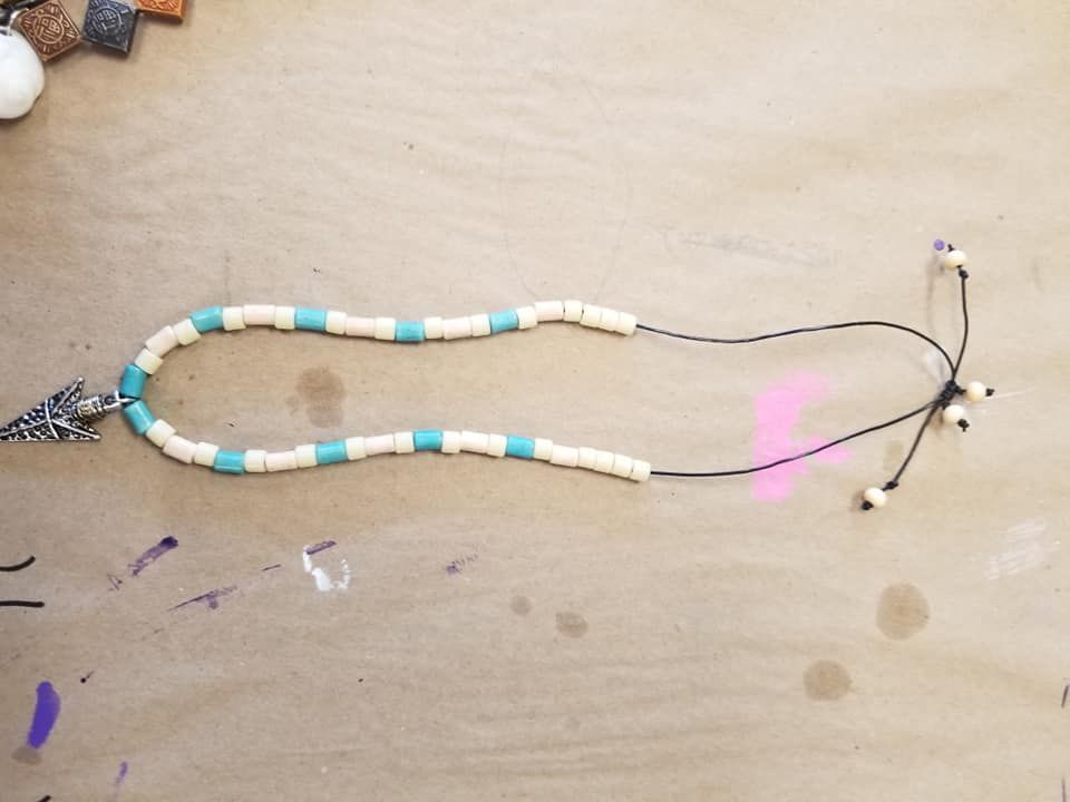 A handmade necklace with white and blue beads, an arrow charm, and adjustable black cord on a craft surface.