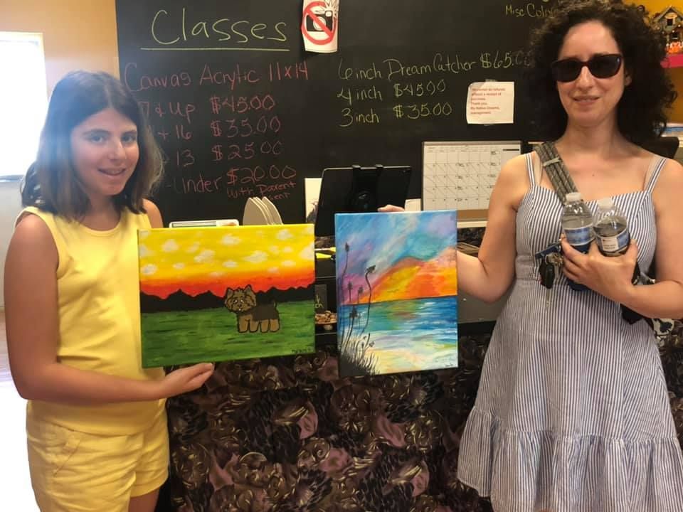 Two people holding paintings in front of a counter. One in yellow, one in blue, both smiling. Sunset paintings.