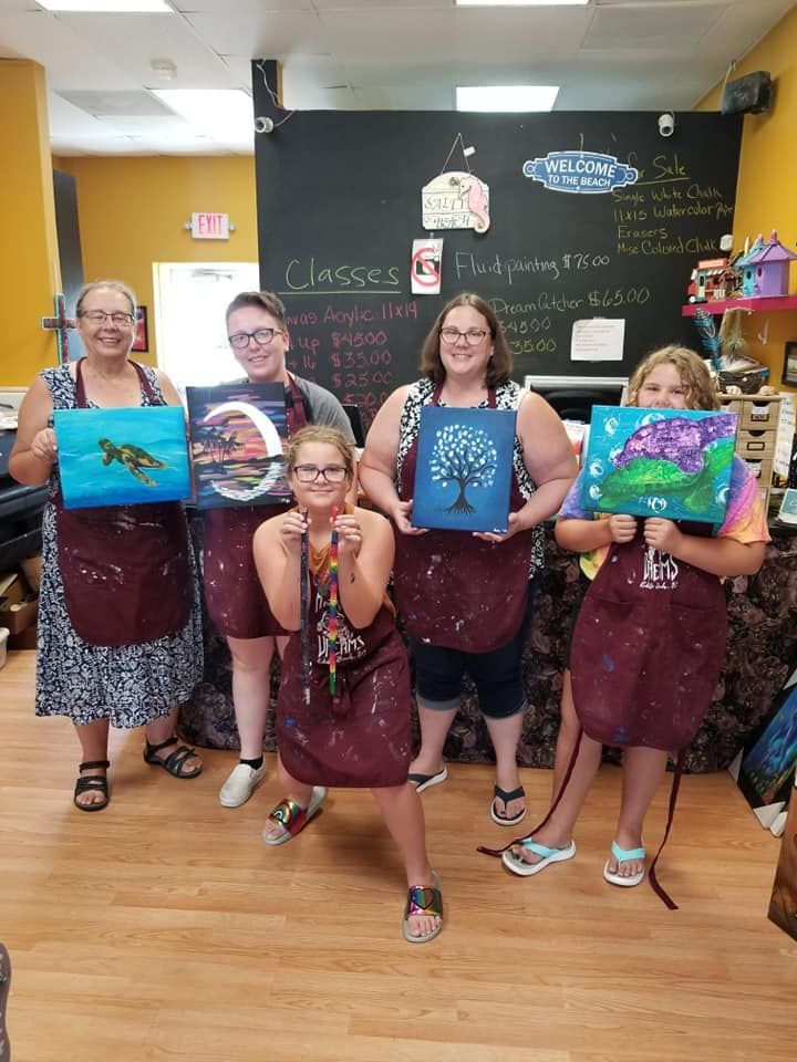 Five people in aprons holding their paintings, smiling in an art class. A blackboard is behind them.