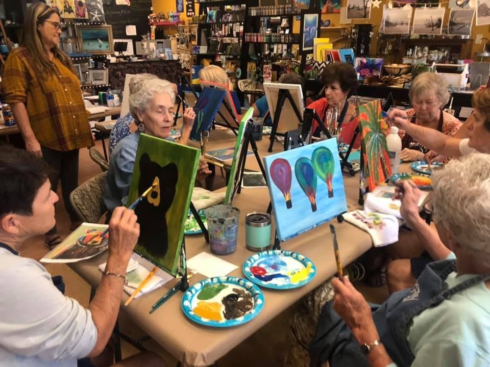 A group of women in a brightly lit art class paint at easels. The instructor looks on.