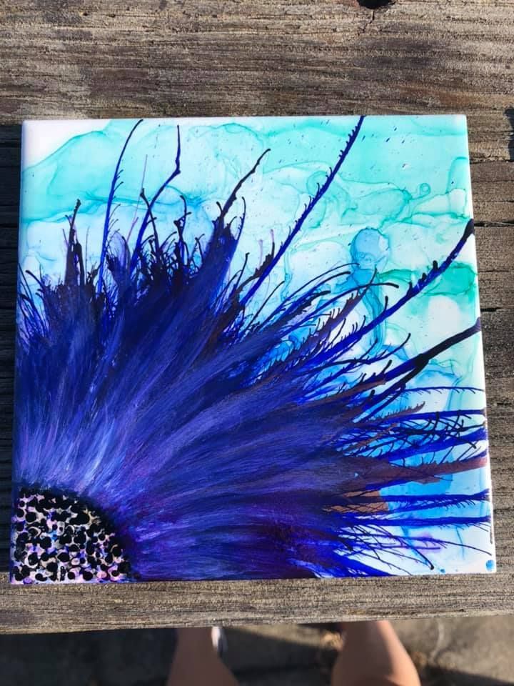 Blue and teal alcohol ink flower on white tile, set outdoors on a wooden surface.