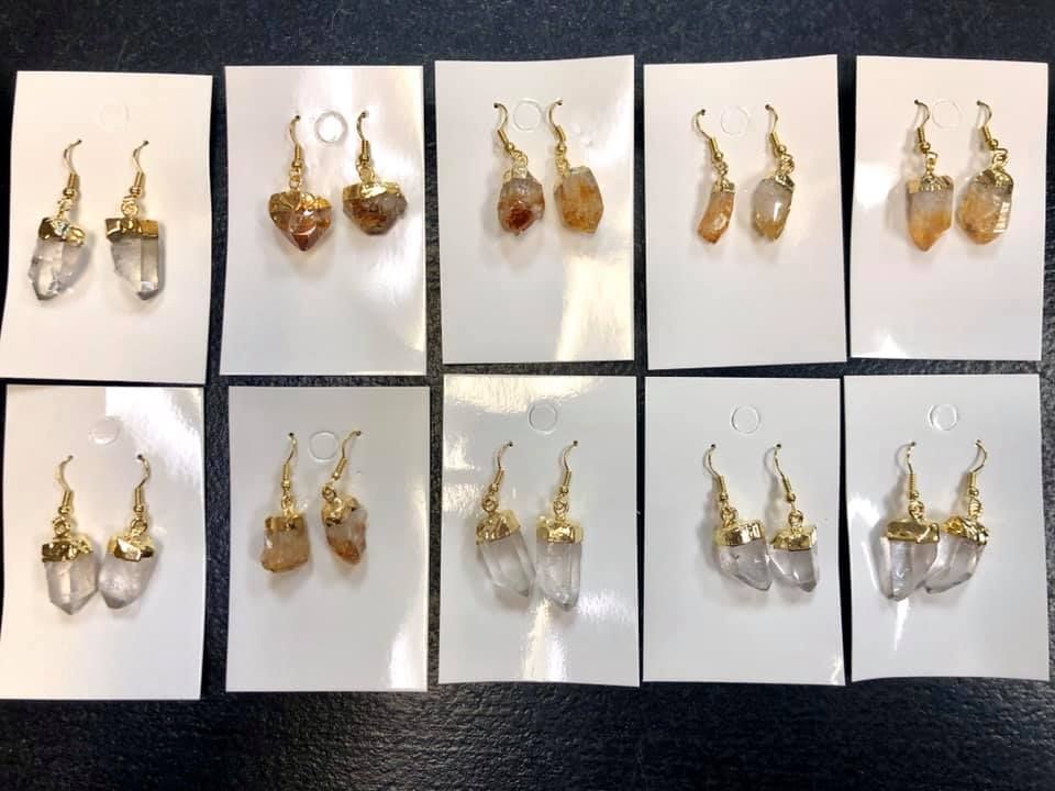 Ten pairs of gold-colored earrings with various crystal or stone pendants displayed on white cards.