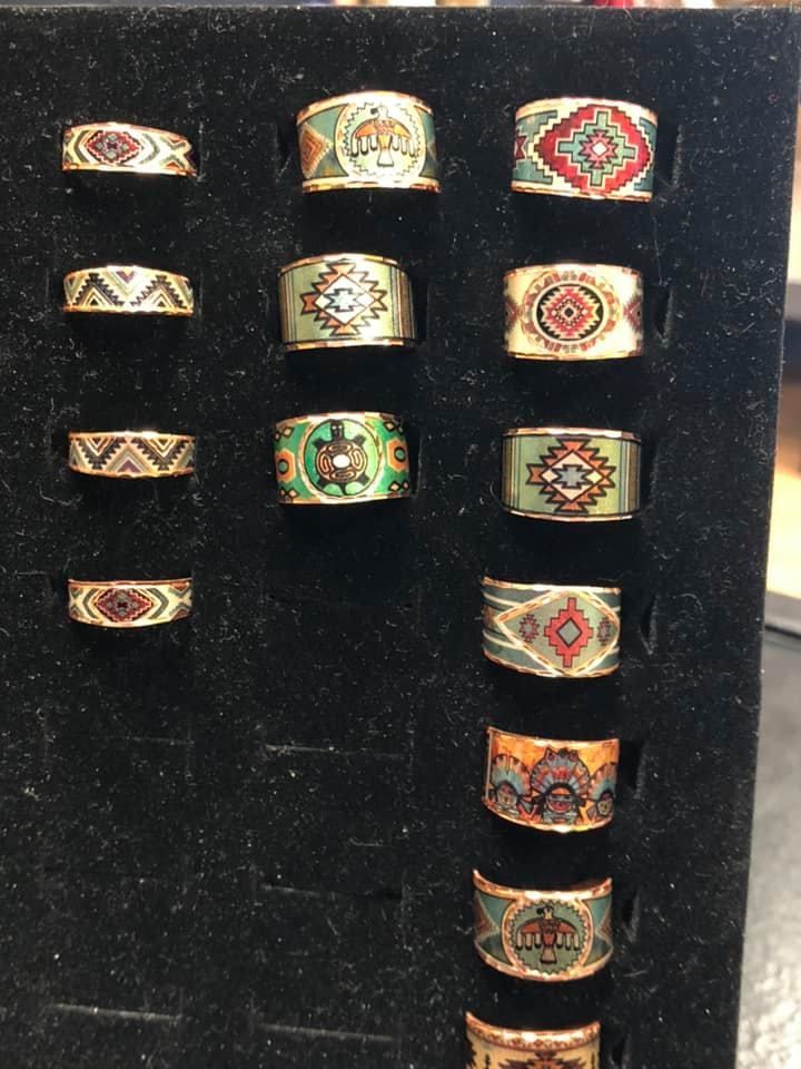 Display of gold-toned rings with colorful, patterned enamel designs. Black display with multiple rings.