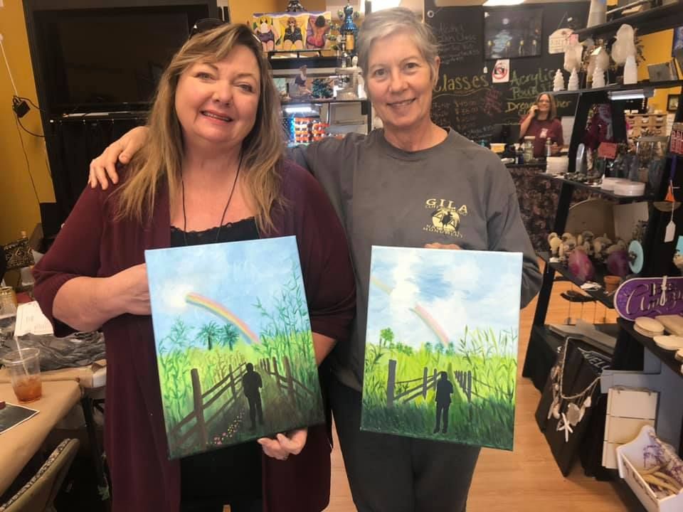 Two women smiling, holding watercolor paintings of a person by a fence under a rainbow.