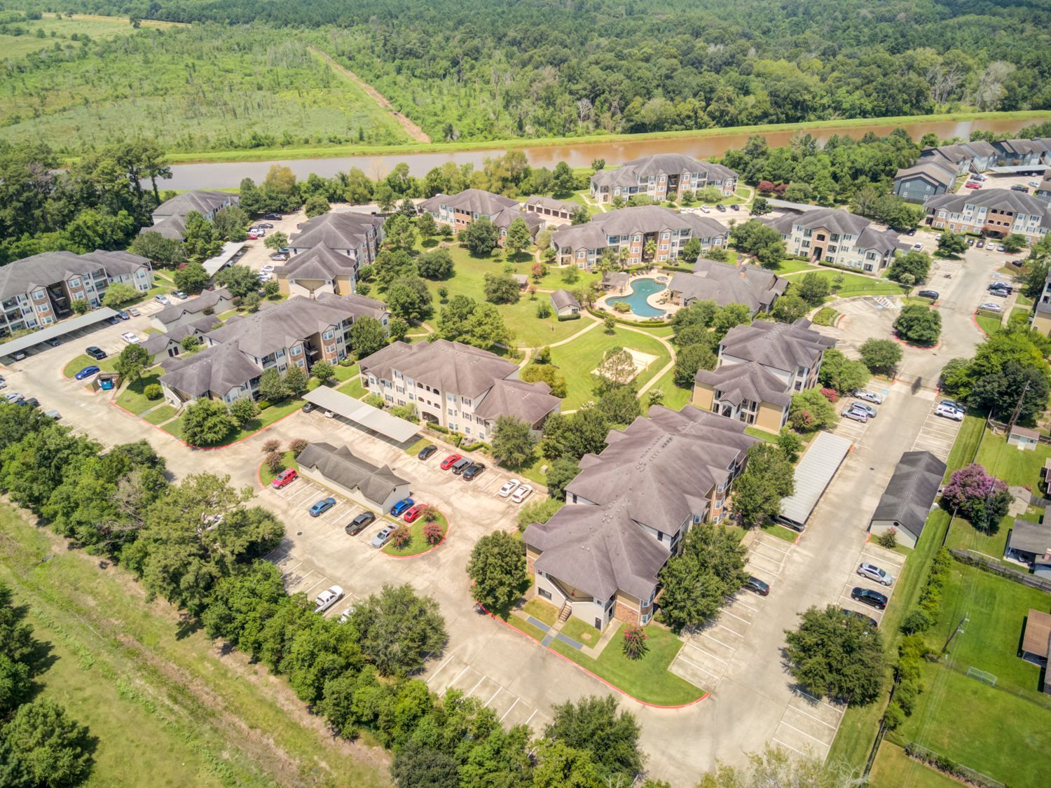 Aerial view of a residential complex with buildings, a pool, trees, and parking; lush greenery in the background.