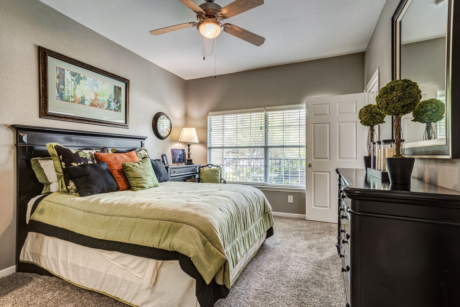 Bedroom with queen bed, desk, dark furniture, green and orange accents, ceiling fan, and window.