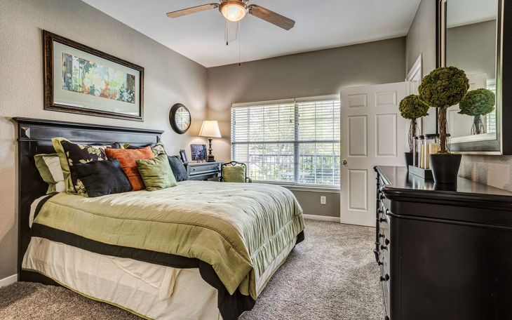 Bedroom with queen bed, desk, dark furniture, green and orange accents, ceiling fan, and window.
