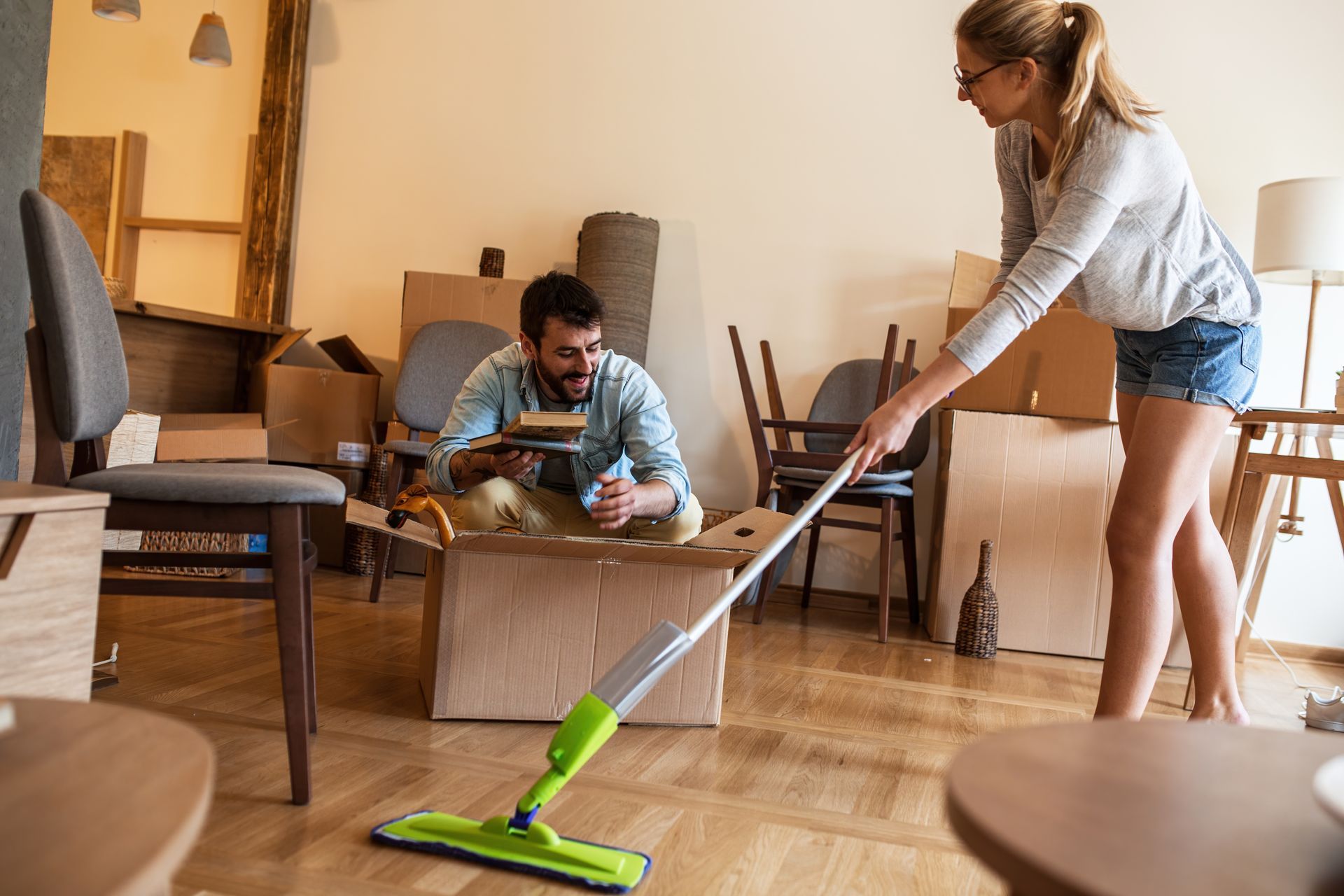 Woman mopping floor, man in cardboard box. Moving boxes and furniture in a room.