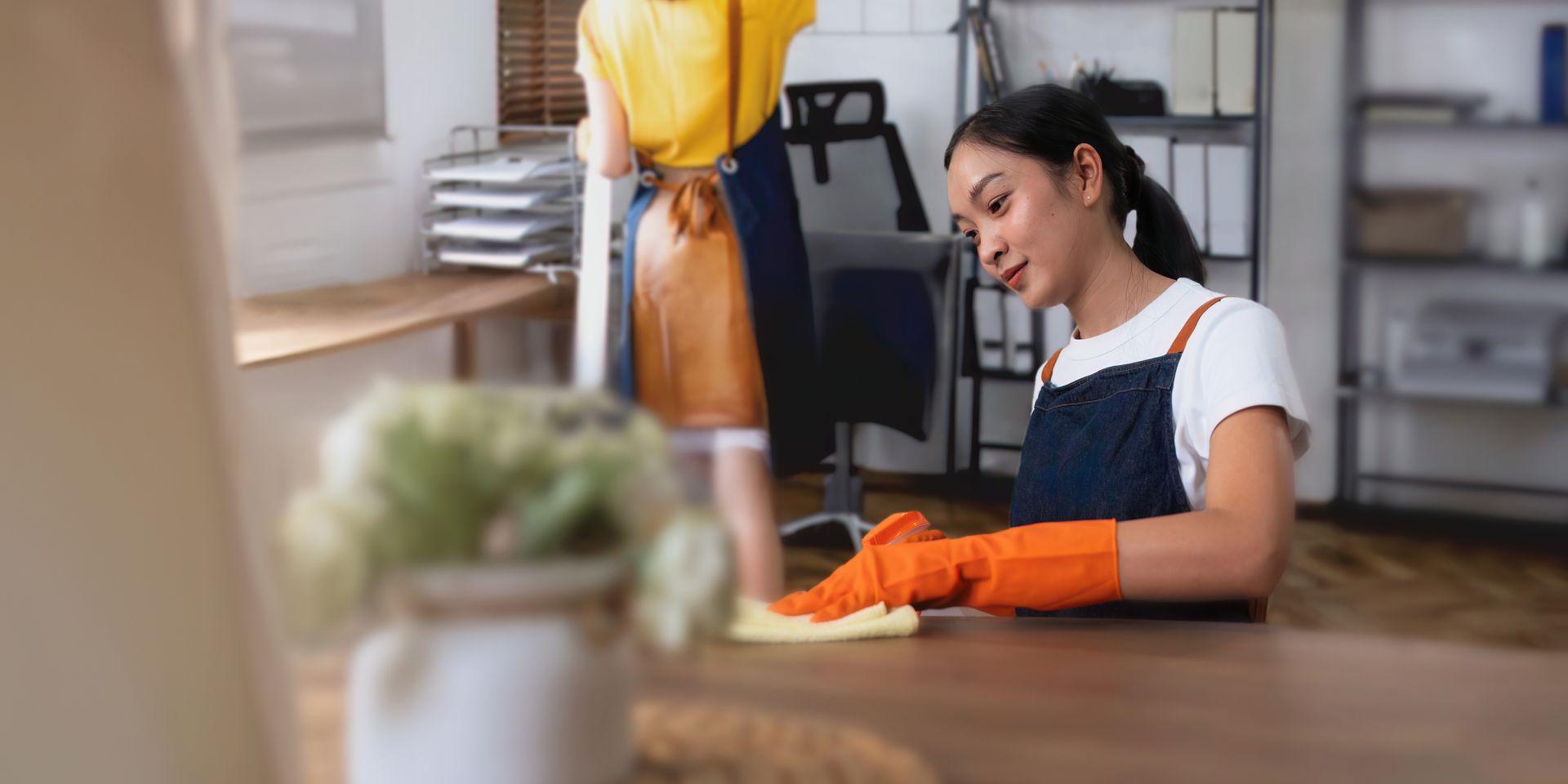 Woman wearing orange gloves, cleaning a wooden surface with a yellow cloth. Another person is in the background.