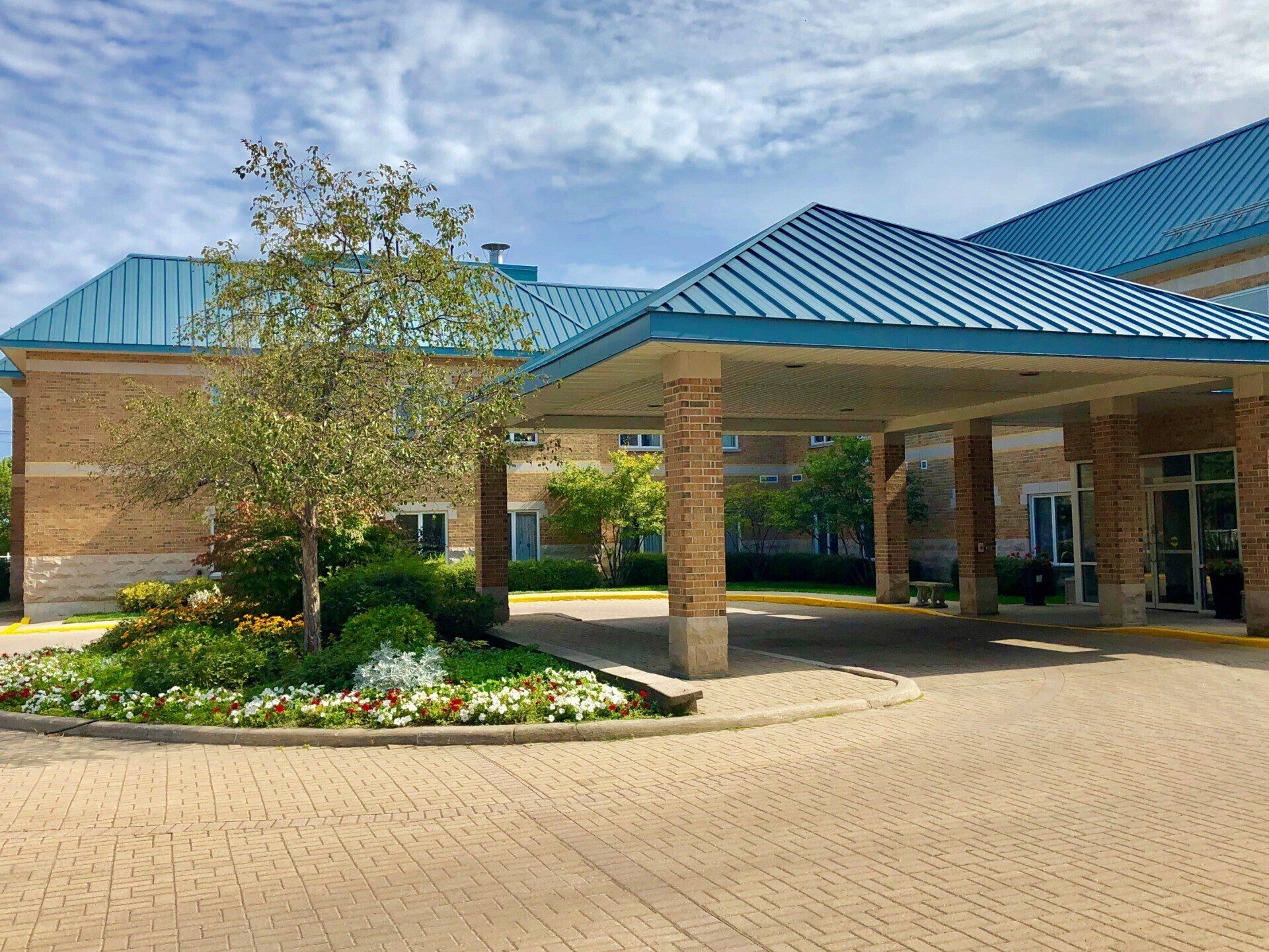 Front entrance to Hillcrest Village Long-Term Care Facility in Midland. A large brick building with a blue roof and a driveway leading to it.