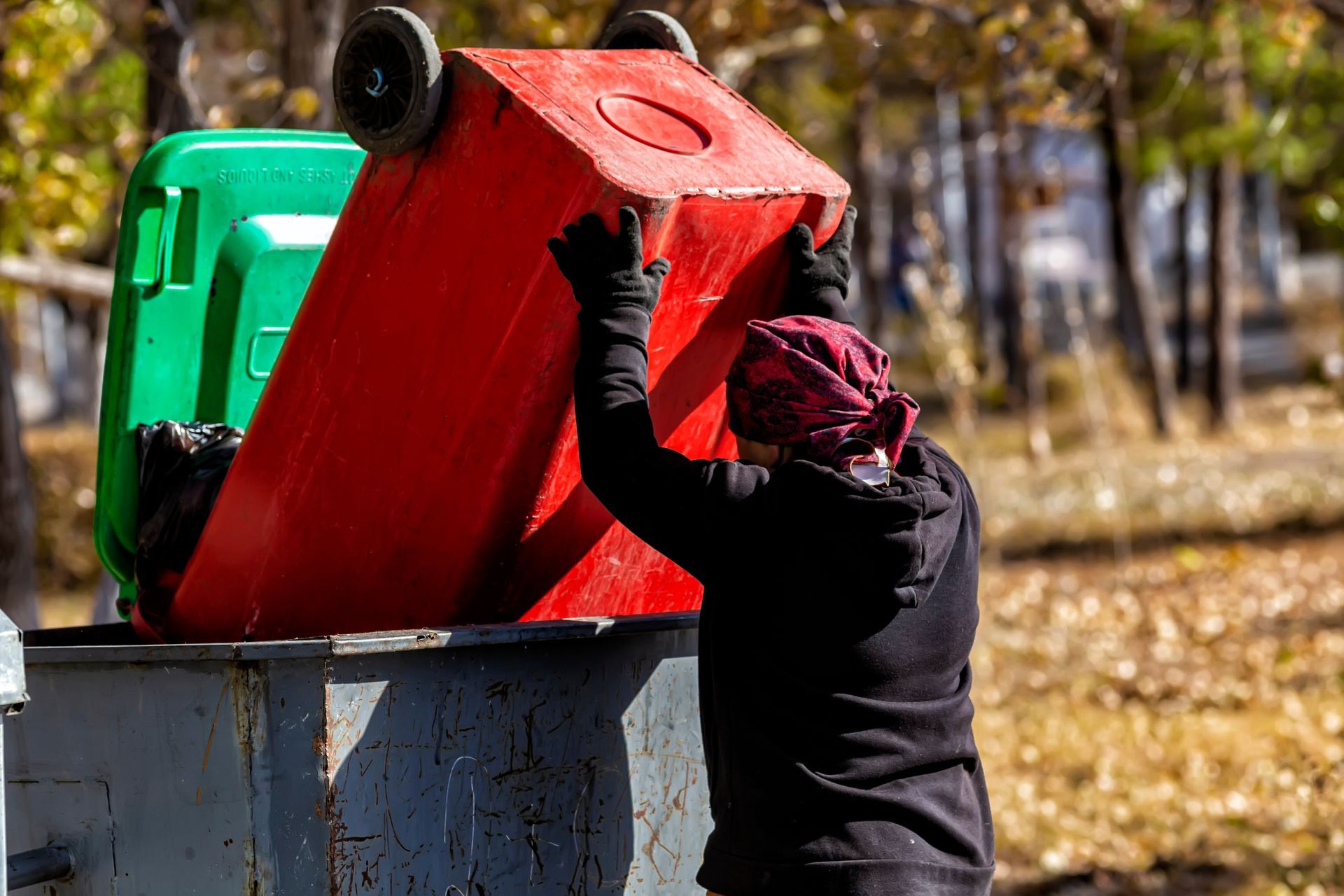 Person emptying a red trash bin into a dumpster. Green trash bin on the left, outdoors.