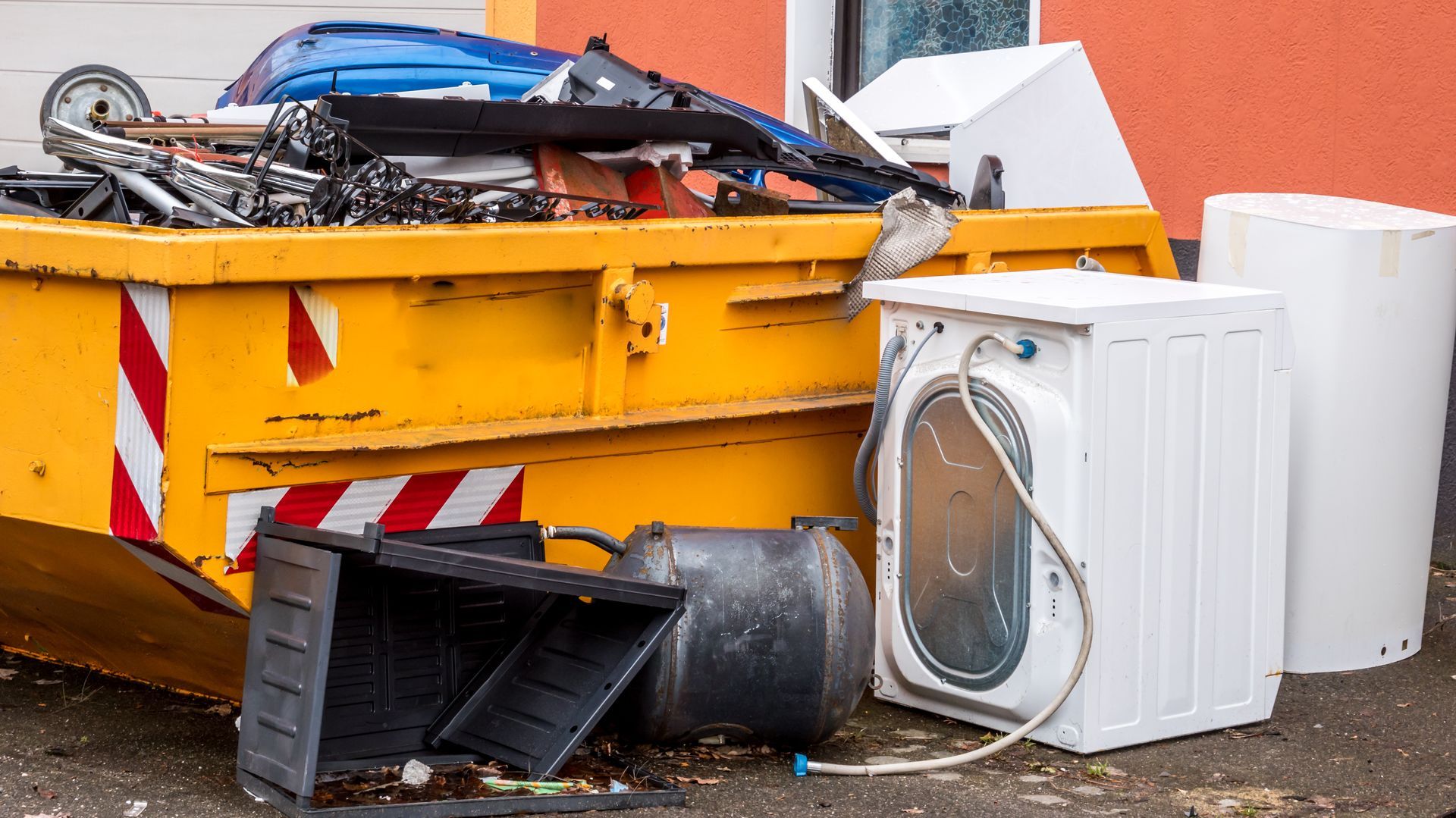 Yellow dumpster overflowing with trash next to a washing machine and other discarded items.