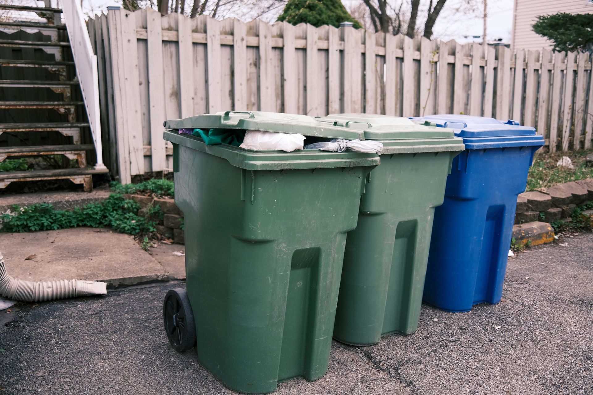 Three trash bins, green and blue, on an asphalt driveway next to a fence and stairs.