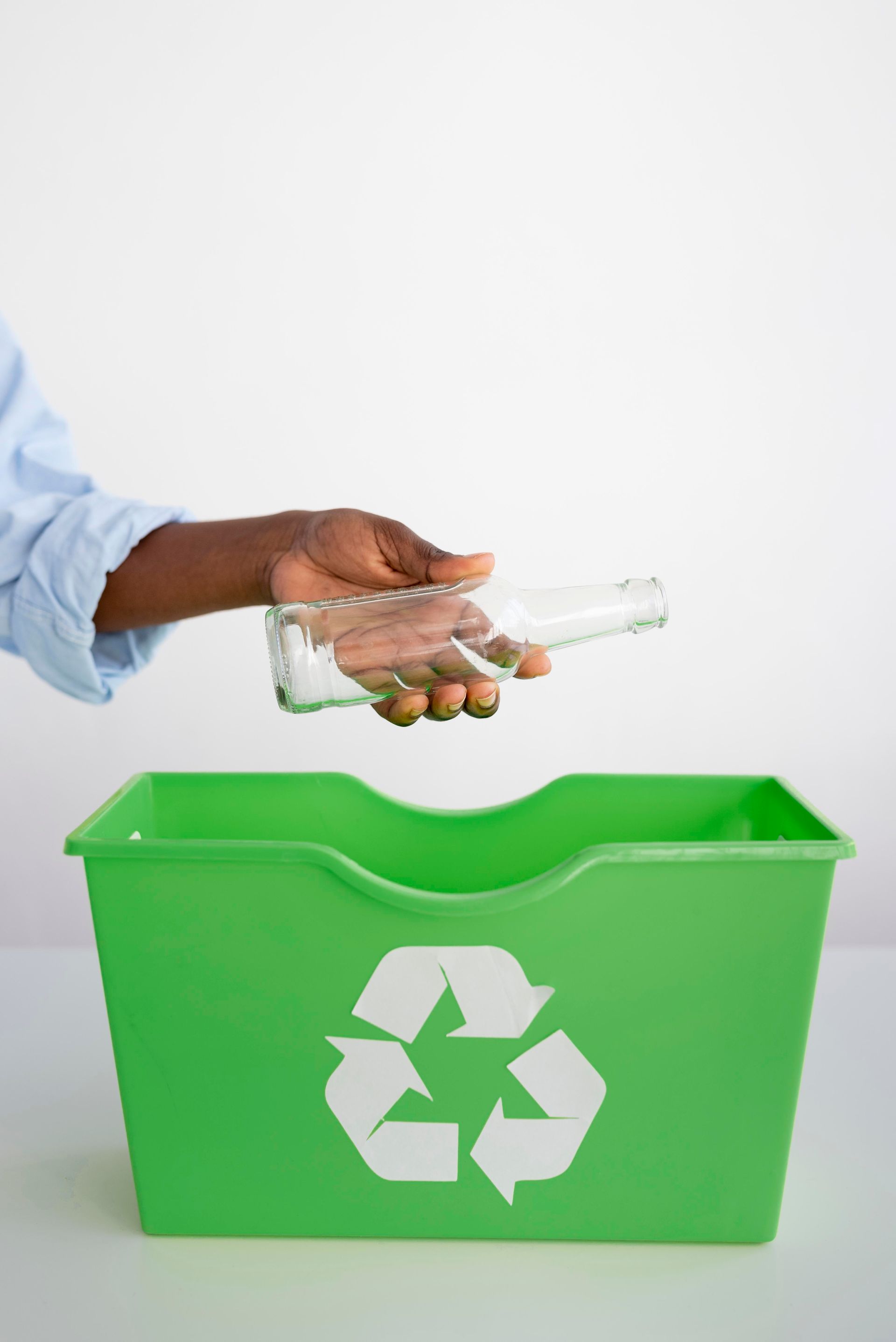A person is putting a glass bottle into a green recycling bin.