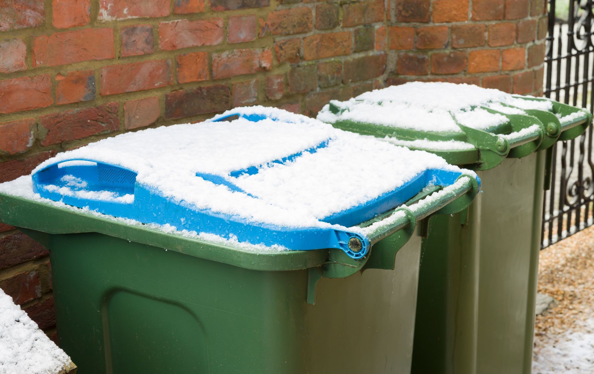 Two green trash bins with snow on their lids, in front of a brick wall and a fence.