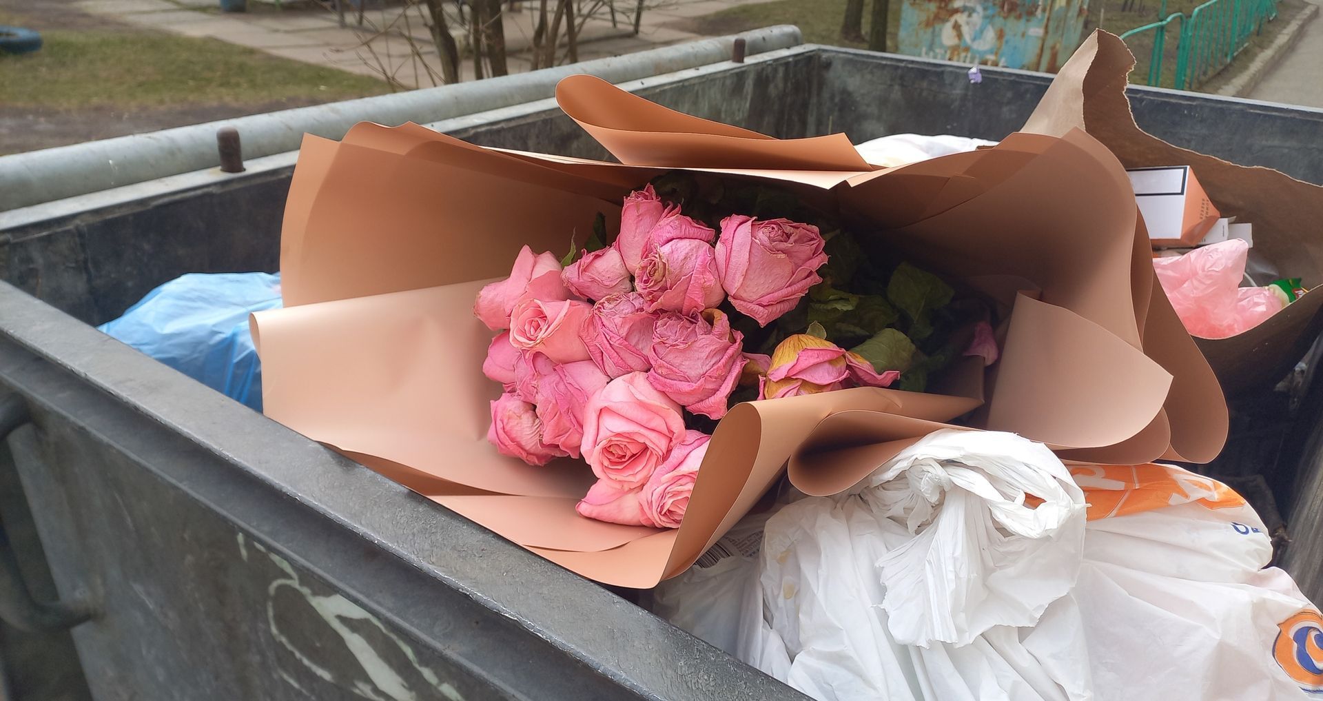 Pink roses in brown paper are discarded in a trash bin.