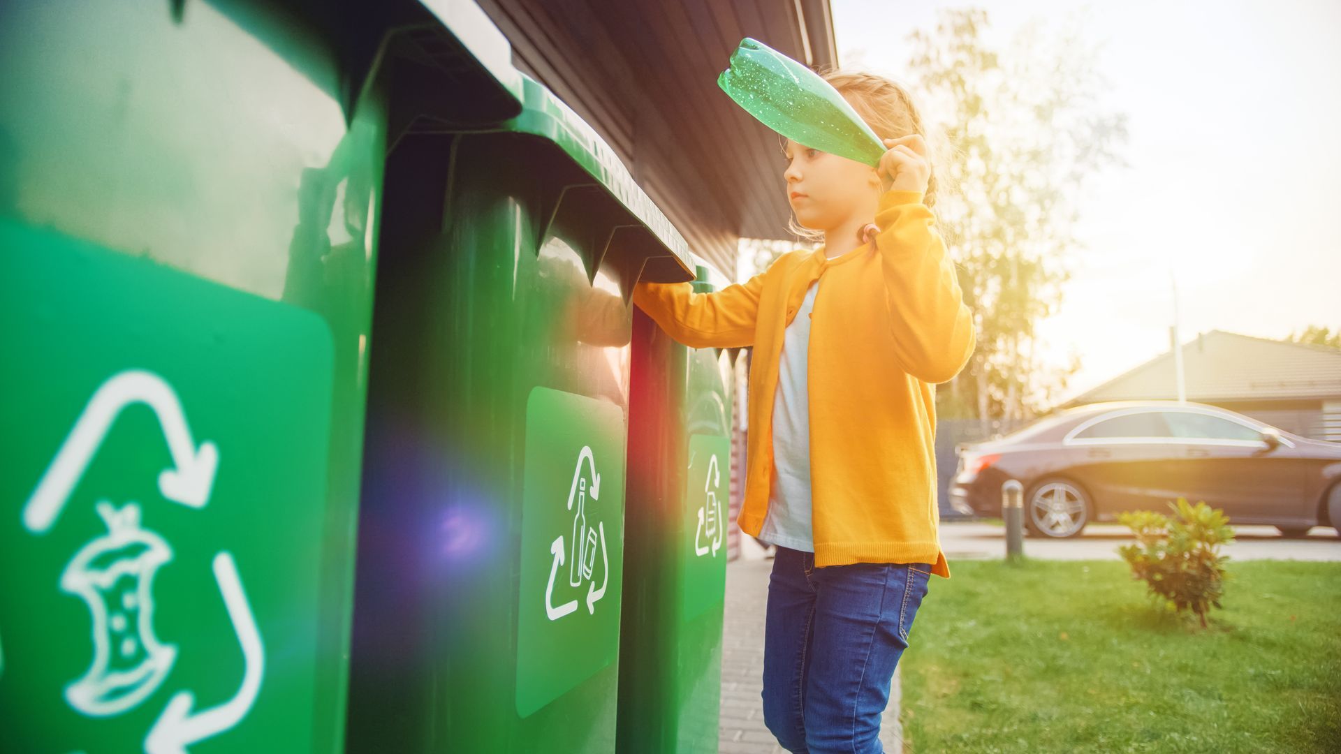 Girl recycling a plastic bottle in a green bin outside on a sunny day.