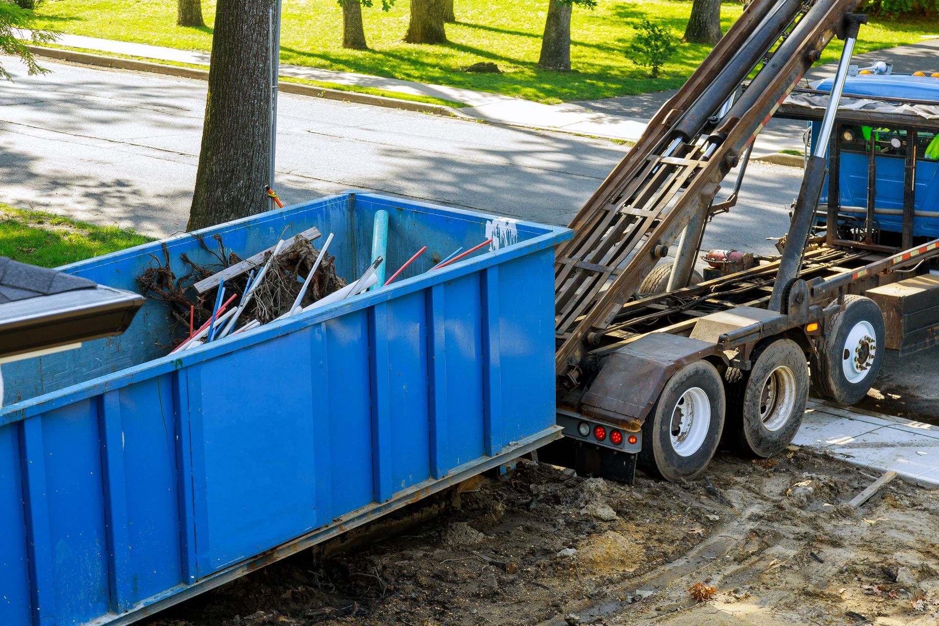 Blue dumpster being lifted by a truck in a residential driveway, filled with debris.