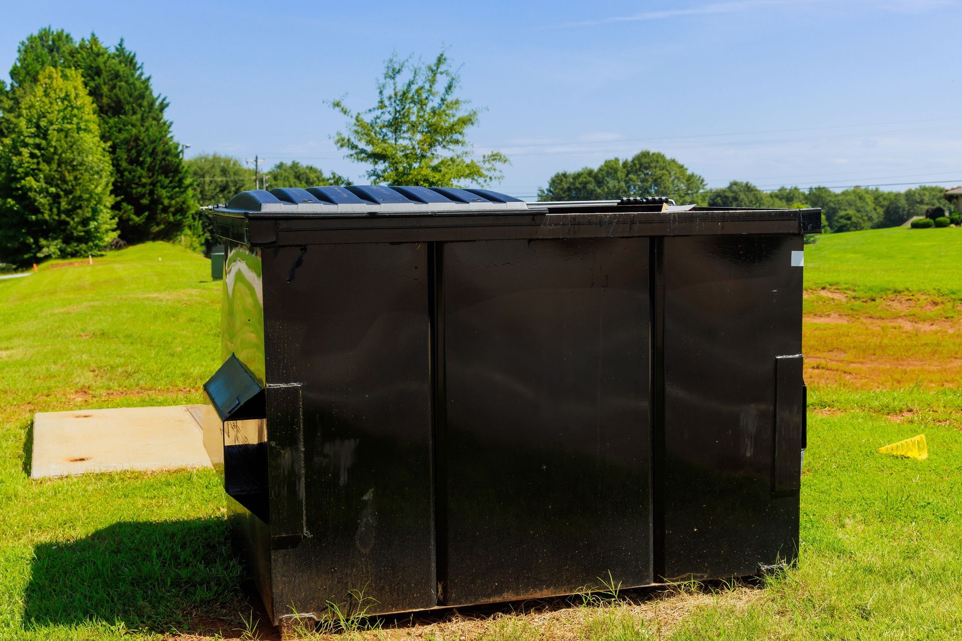 Black dumpster on green grass with trees and blue sky.