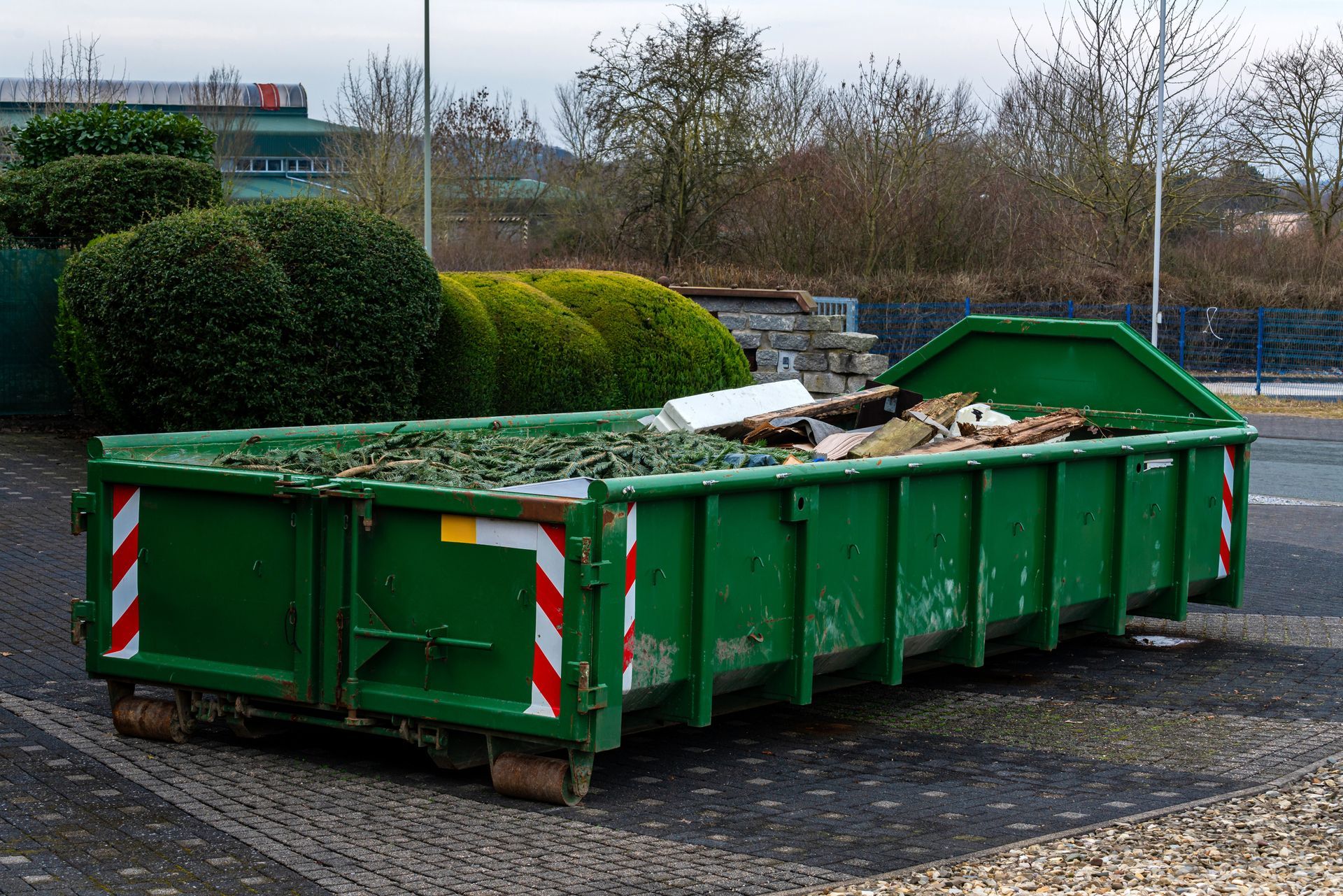 Green waste dumpster filled with debris, outdoors near landscaping.