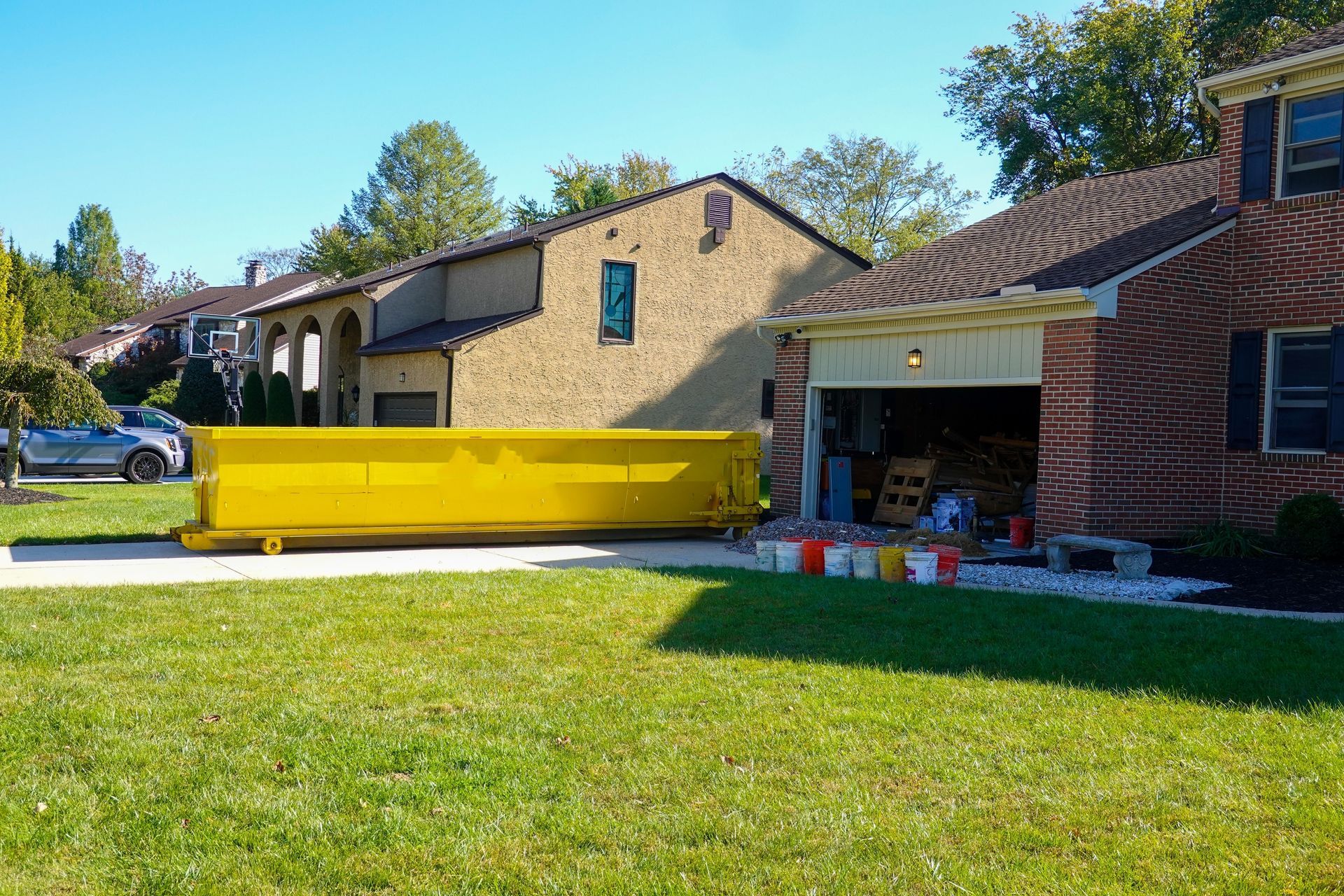 Yellow dumpster in a driveway next to a brick house and an open garage with paint supplies.