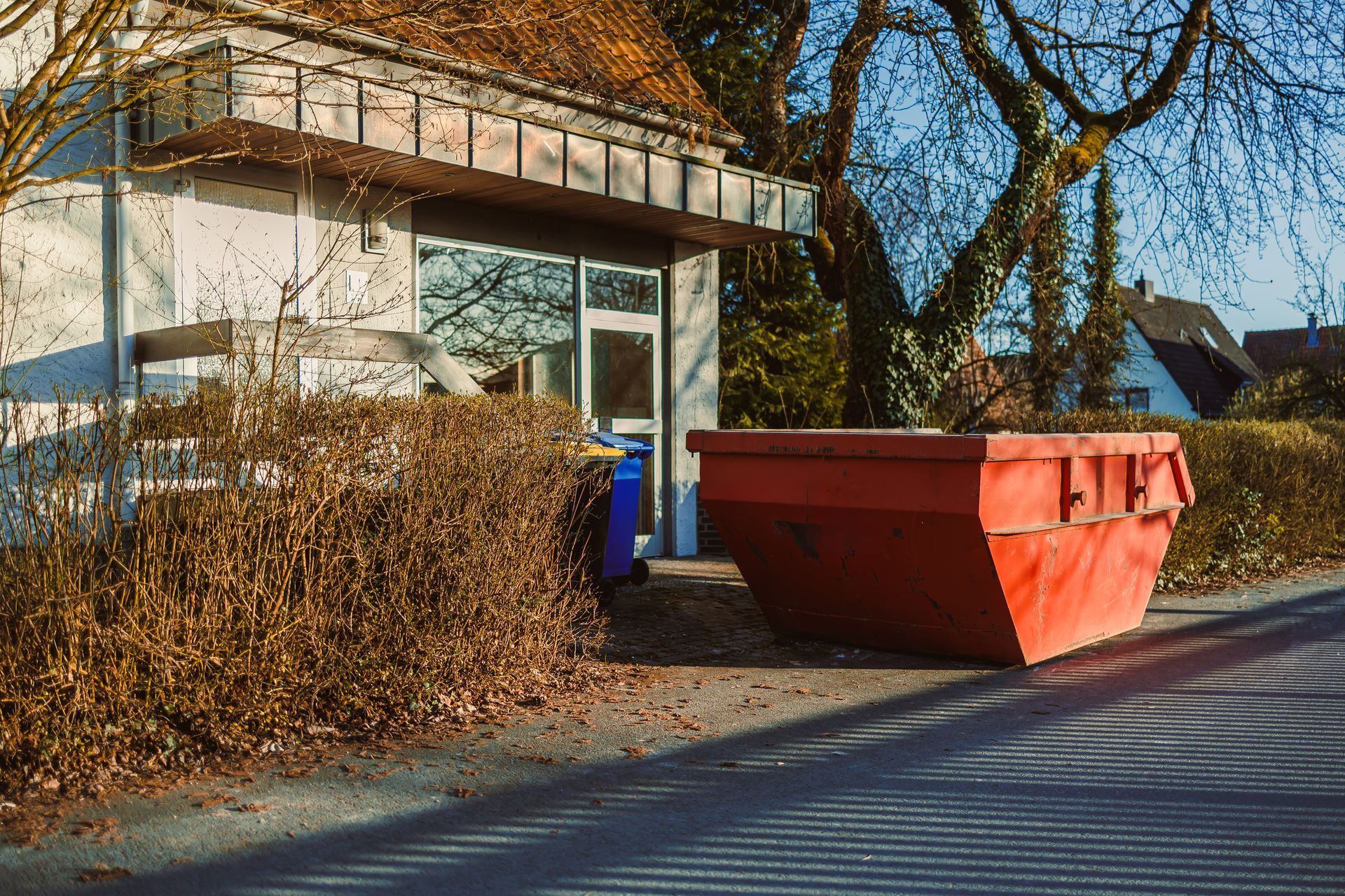A large red industrial dumpster sits on a paved area in front of a building and hedge, next to a smaller blue bin.