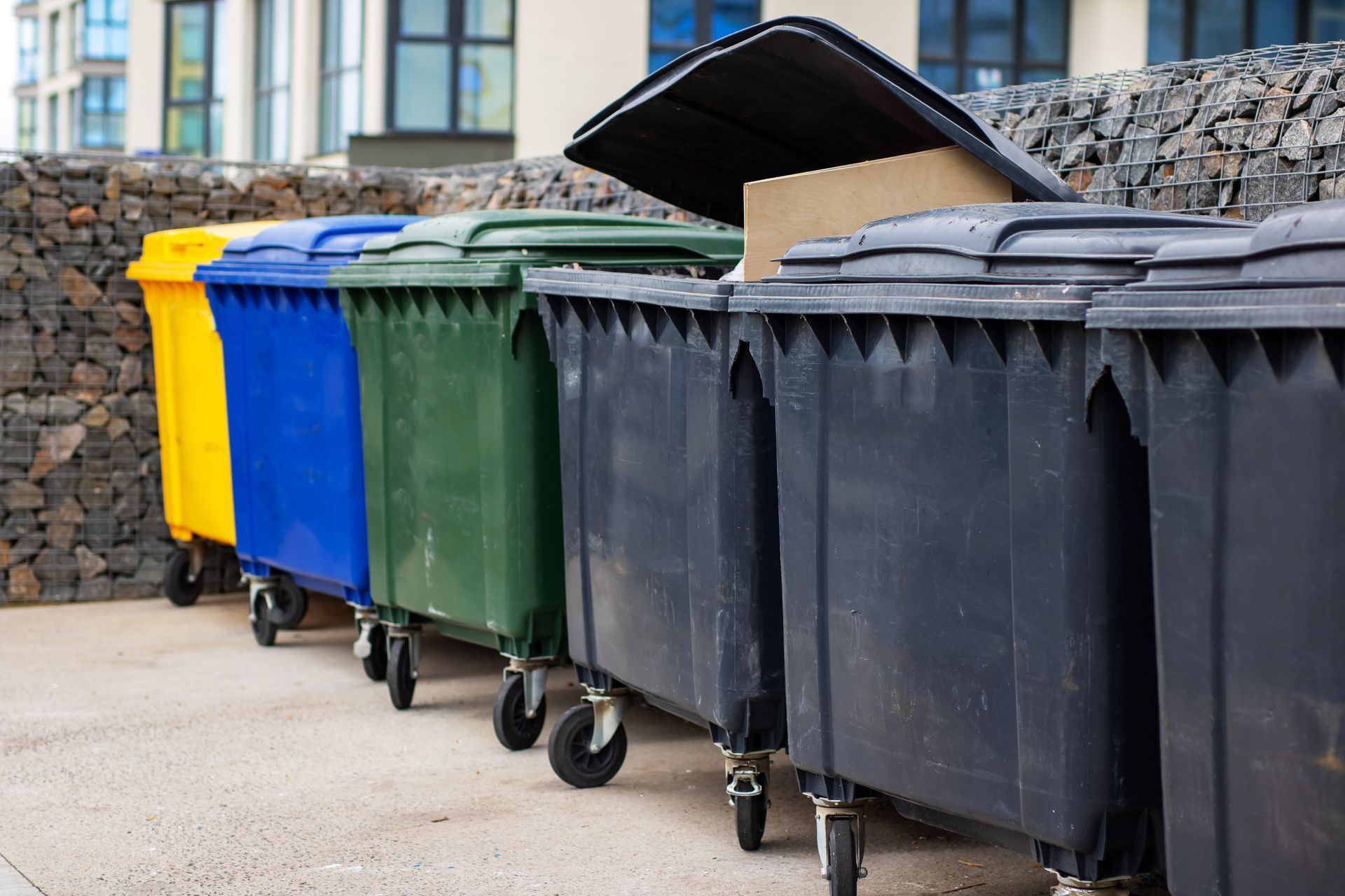 A row of yellow, blue, green, and black dumpsters standing on a paved area against a stone retaining wall.