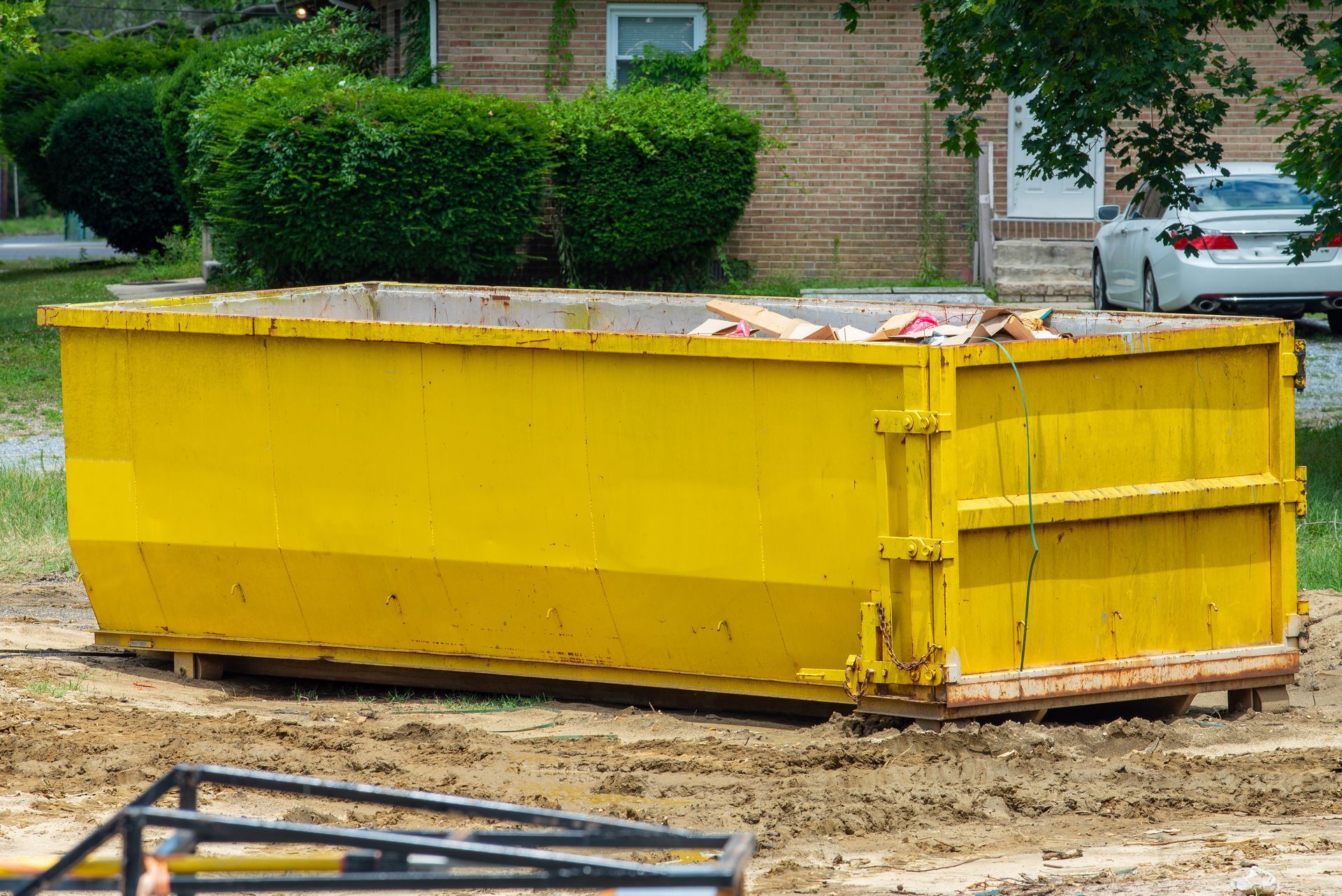 A bright yellow roll-off dumpster sits on a patch of dirt in front of a residential brick house.