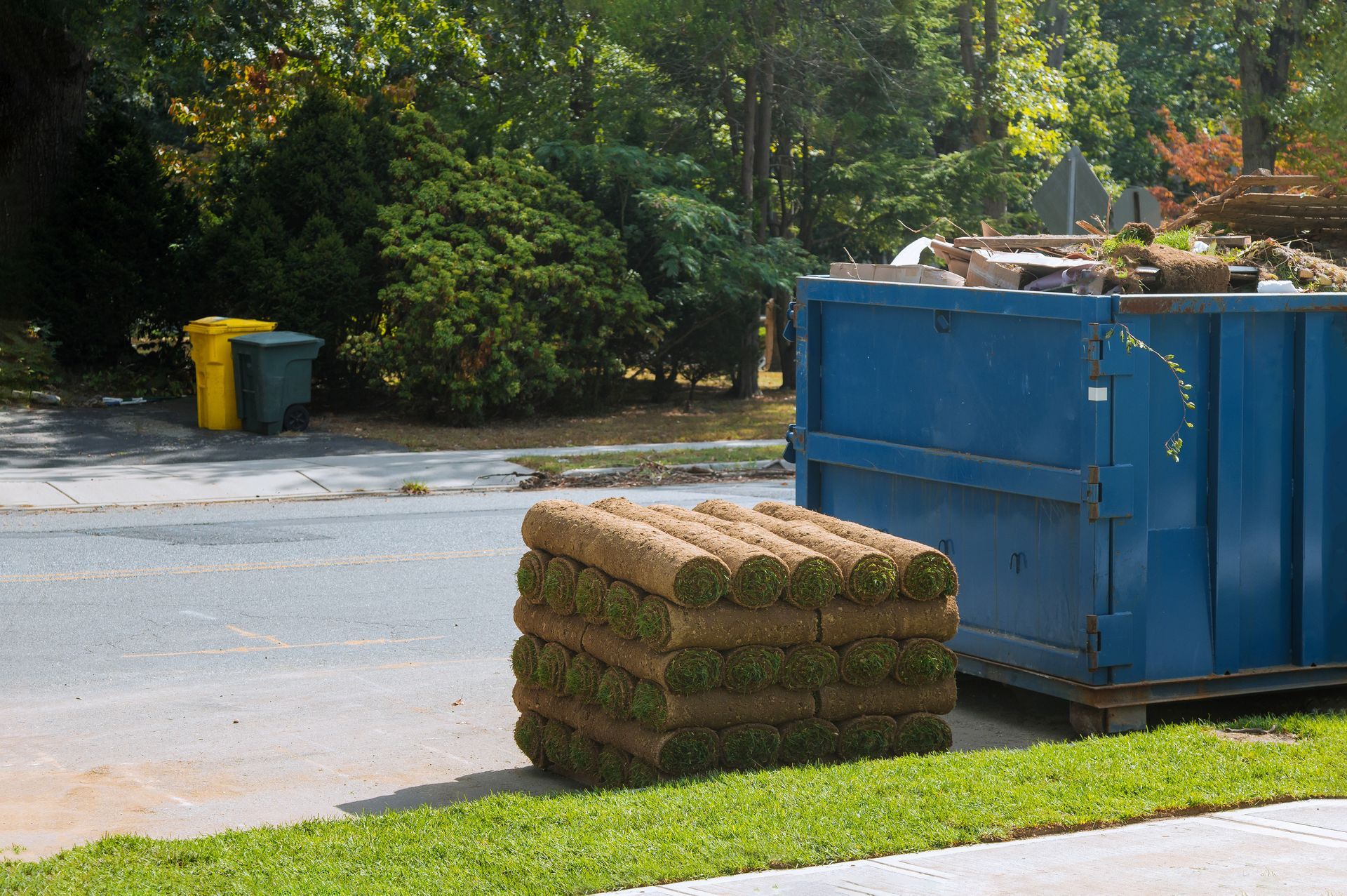 Stacked rolls of sod sit on a paved driveway next to a large blue dumpster, with trash cans and trees in the background.