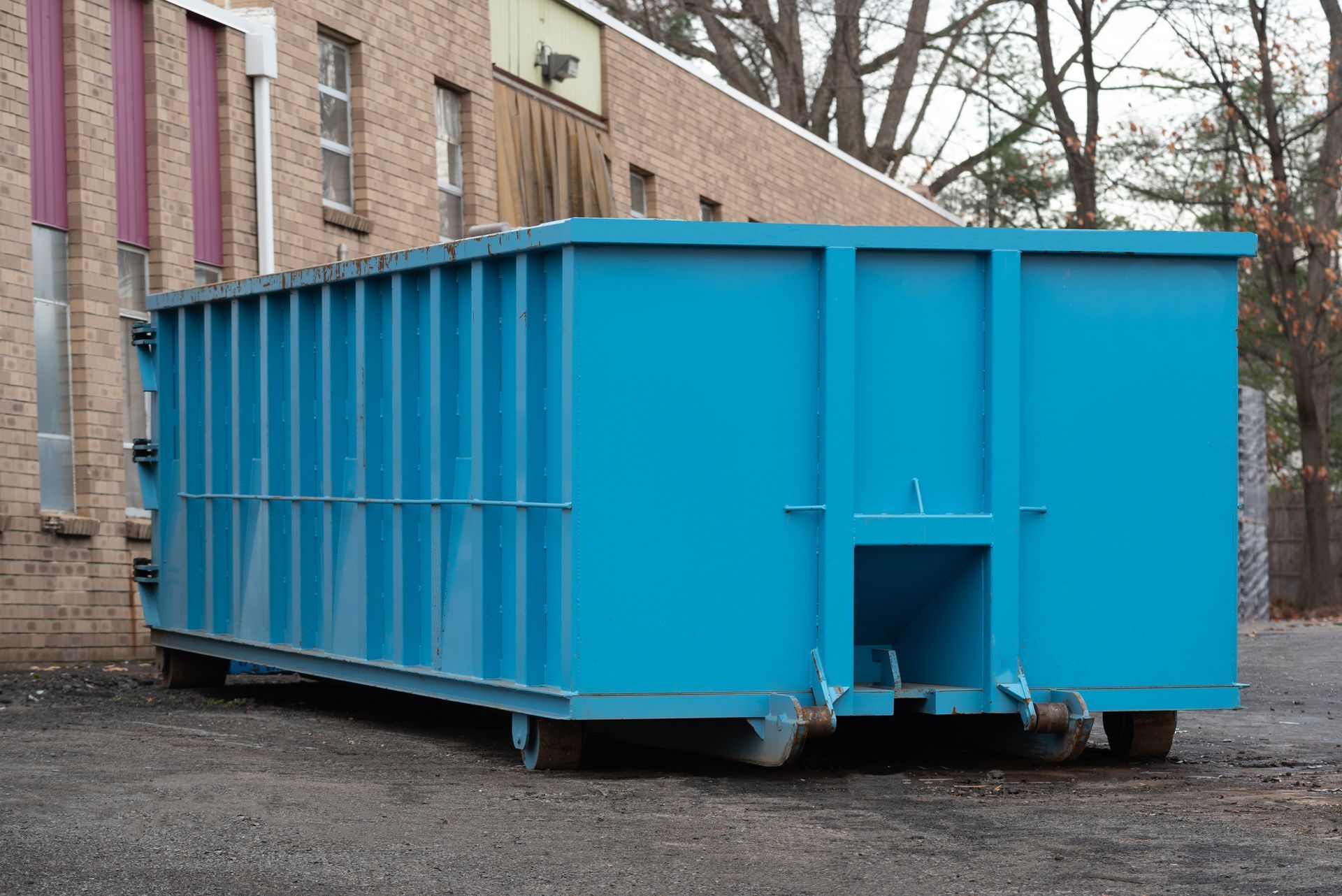 Large blue dumpster against a brick building on a gravel surface.