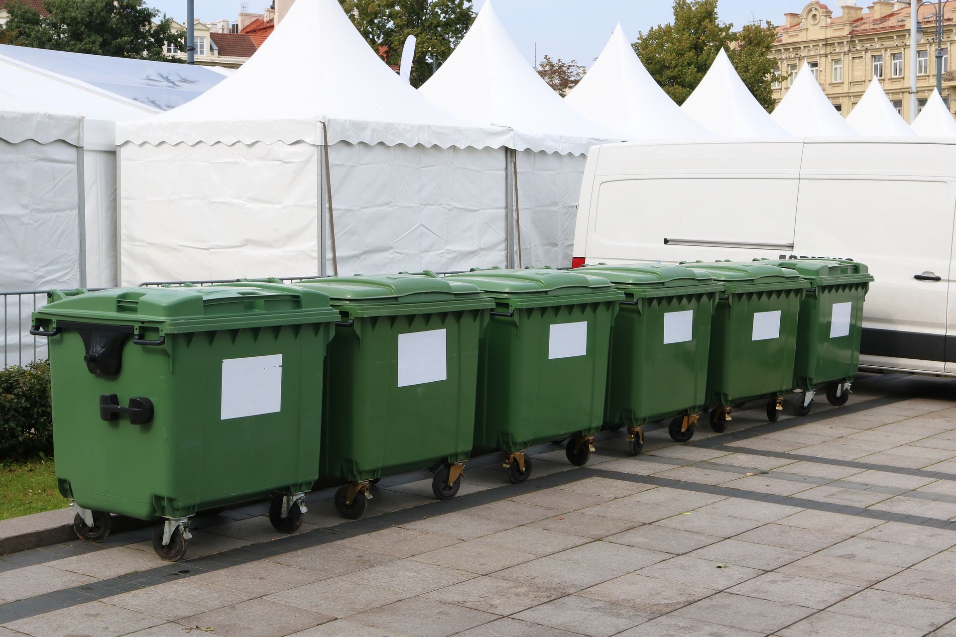 Green garbage bins lined up on pavement, white tents in the background.