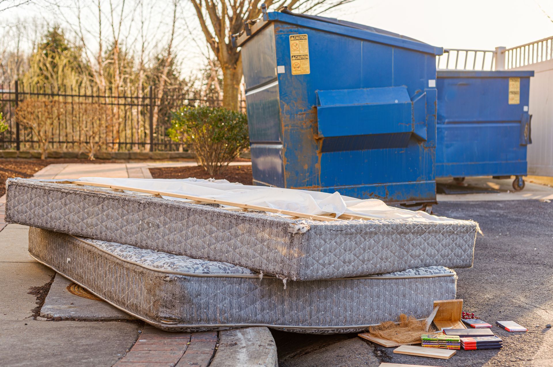 Two discarded mattresses stacked on a sidewalk next to blue dumpsters in an outdoor setting.