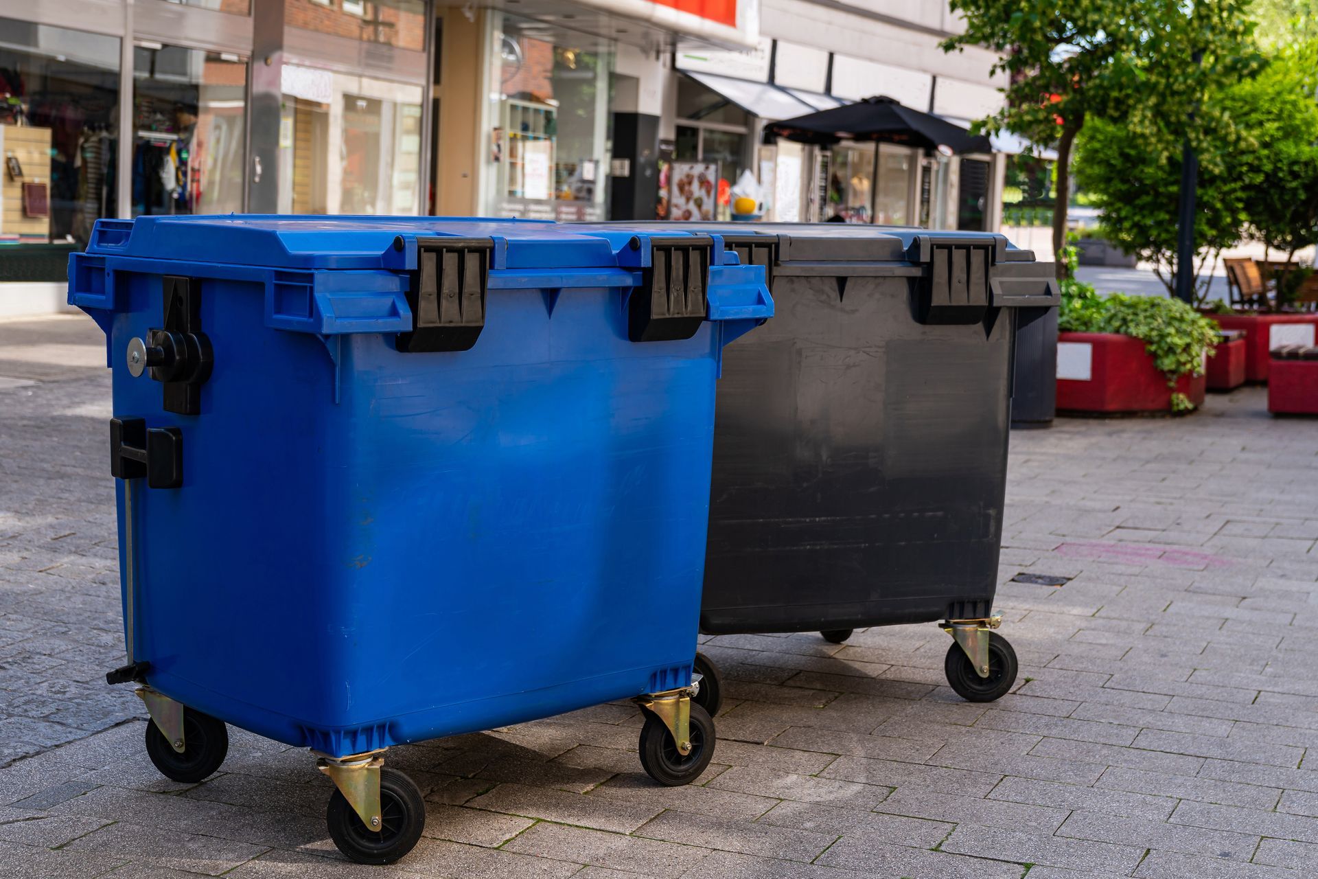 A blue and a black plastic dumpster with wheels sit side-by-side on a paved city sidewalk.