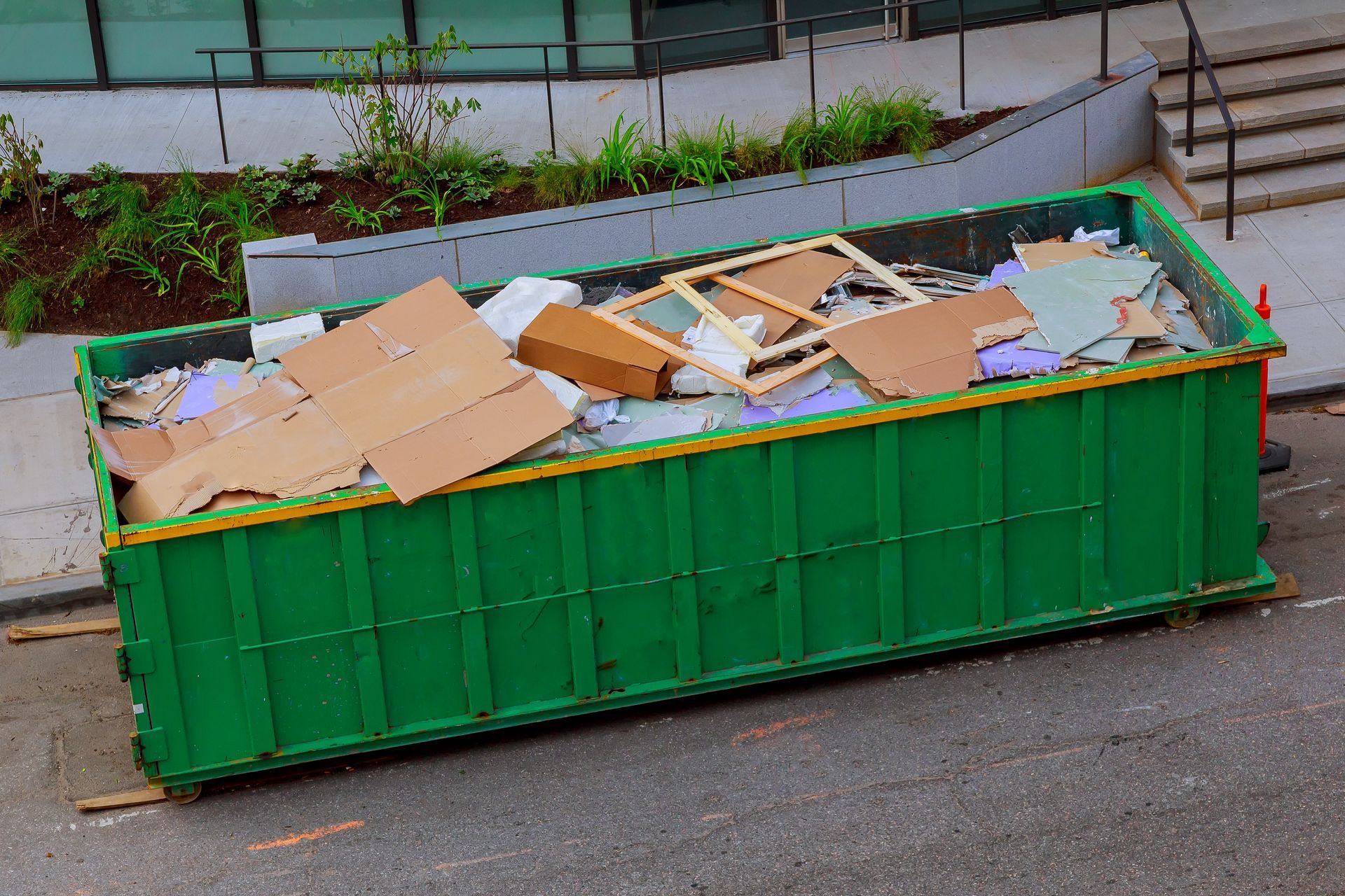 A large, rectangular green dumpster filled with cardboard boxes and construction debris sits on an asphalt street.