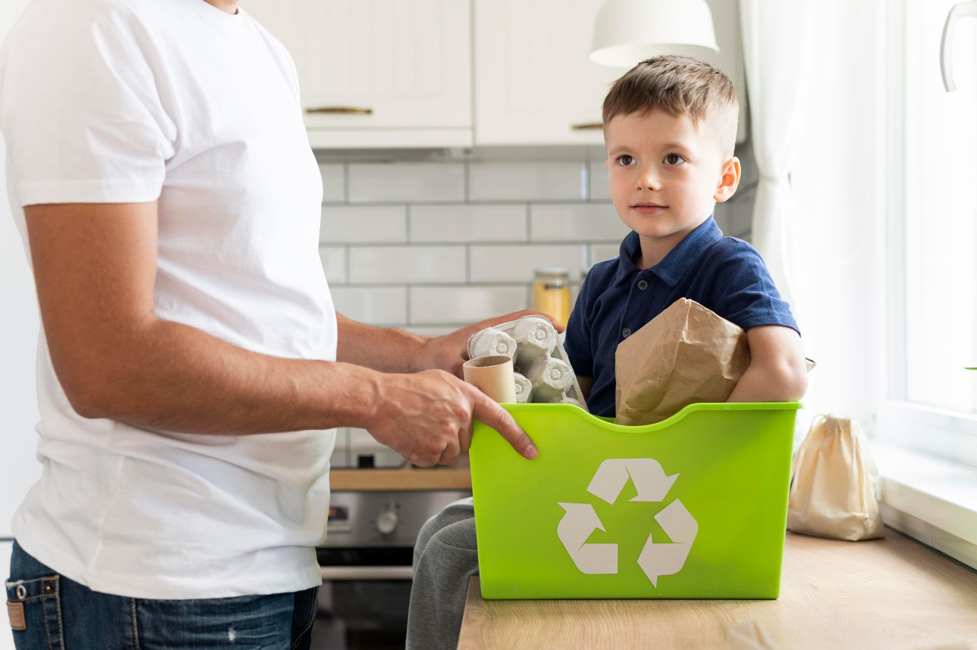 A man and a boy are putting recycling into a green recycling bin.