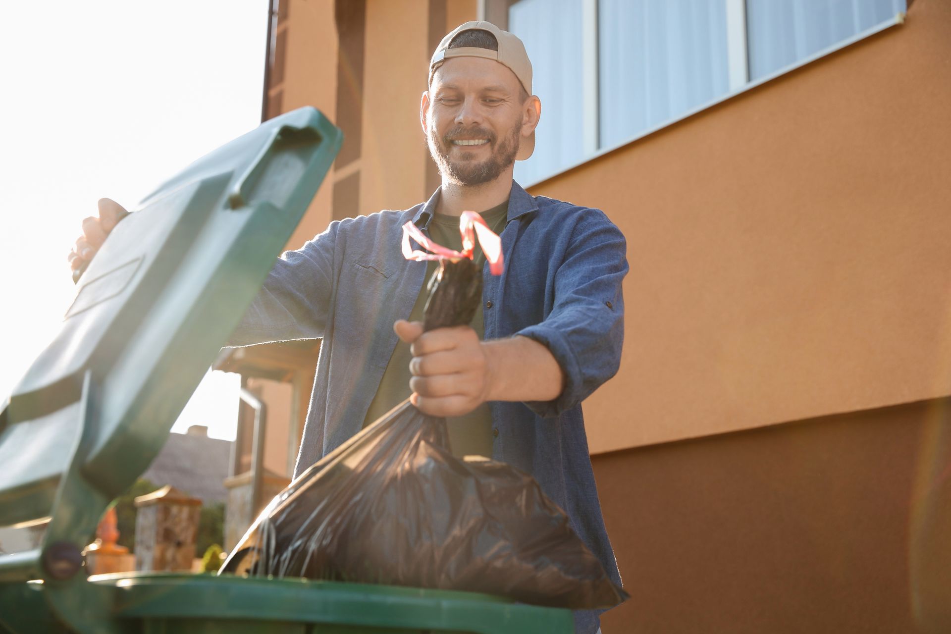 Man emptying a trash bag into a green bin outside a house, smiling.