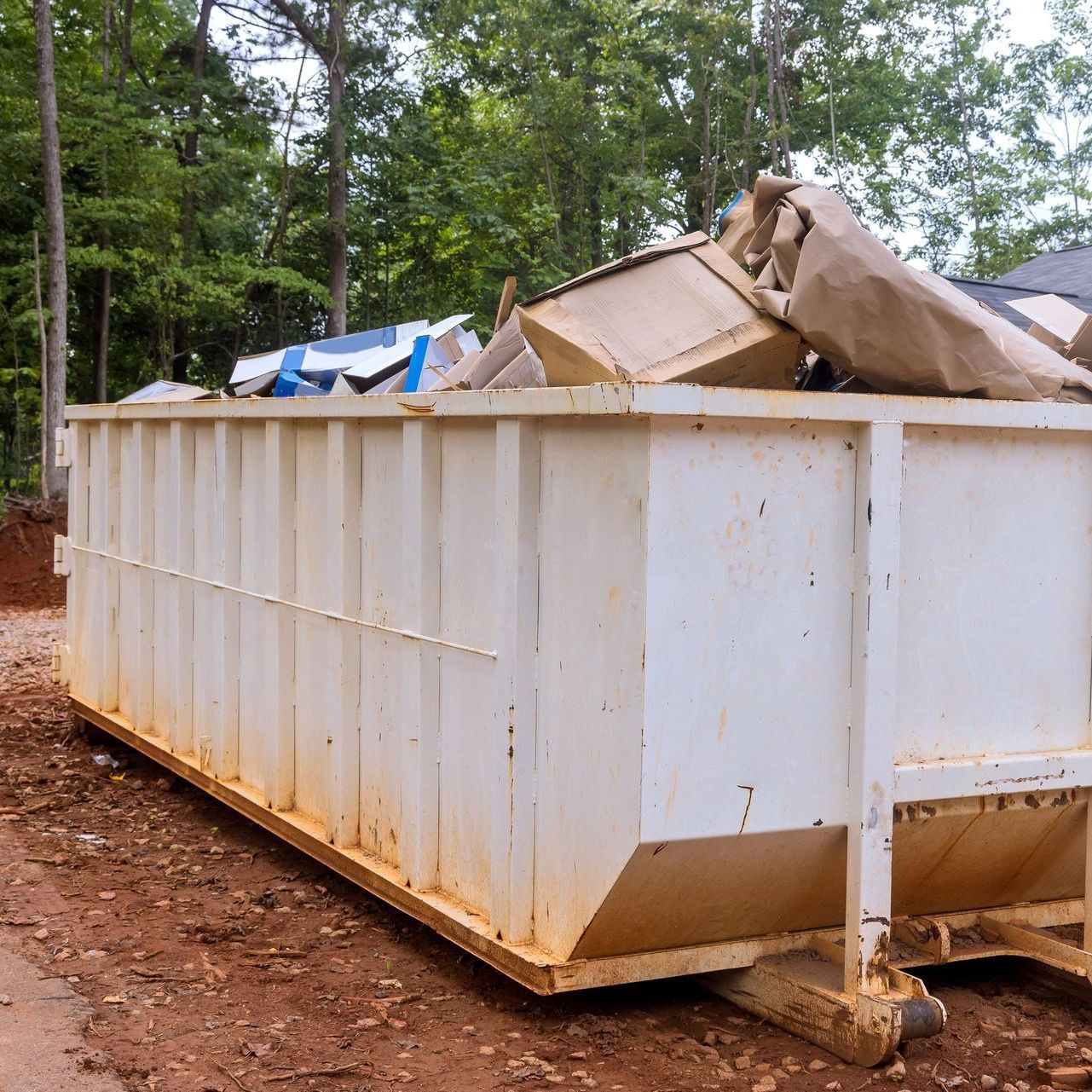 A large white dumpster filled with garbage is sitting on the side of a dirt road.