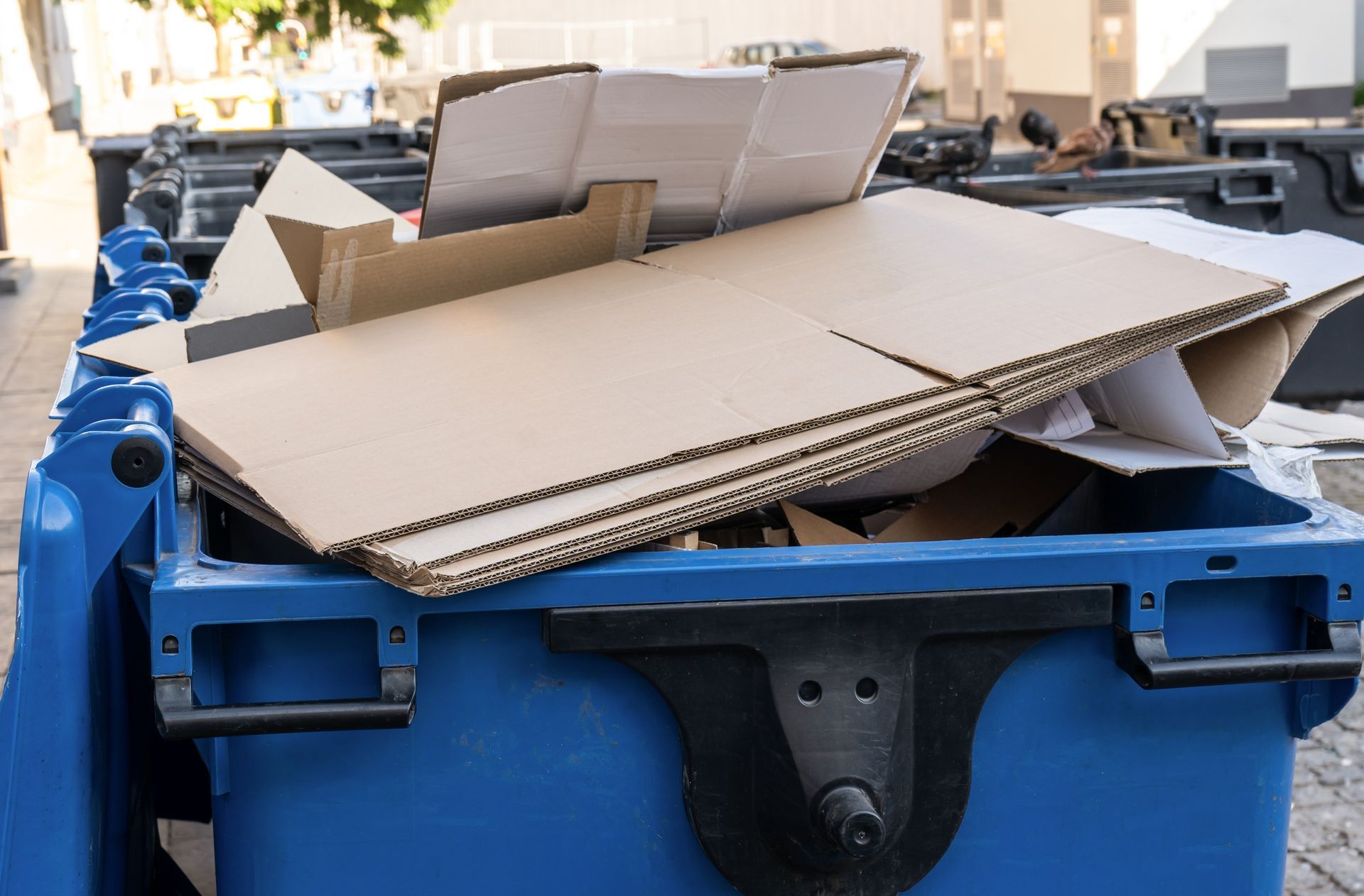 A blue dumpster filled with cardboard boxes on the sidewalk.