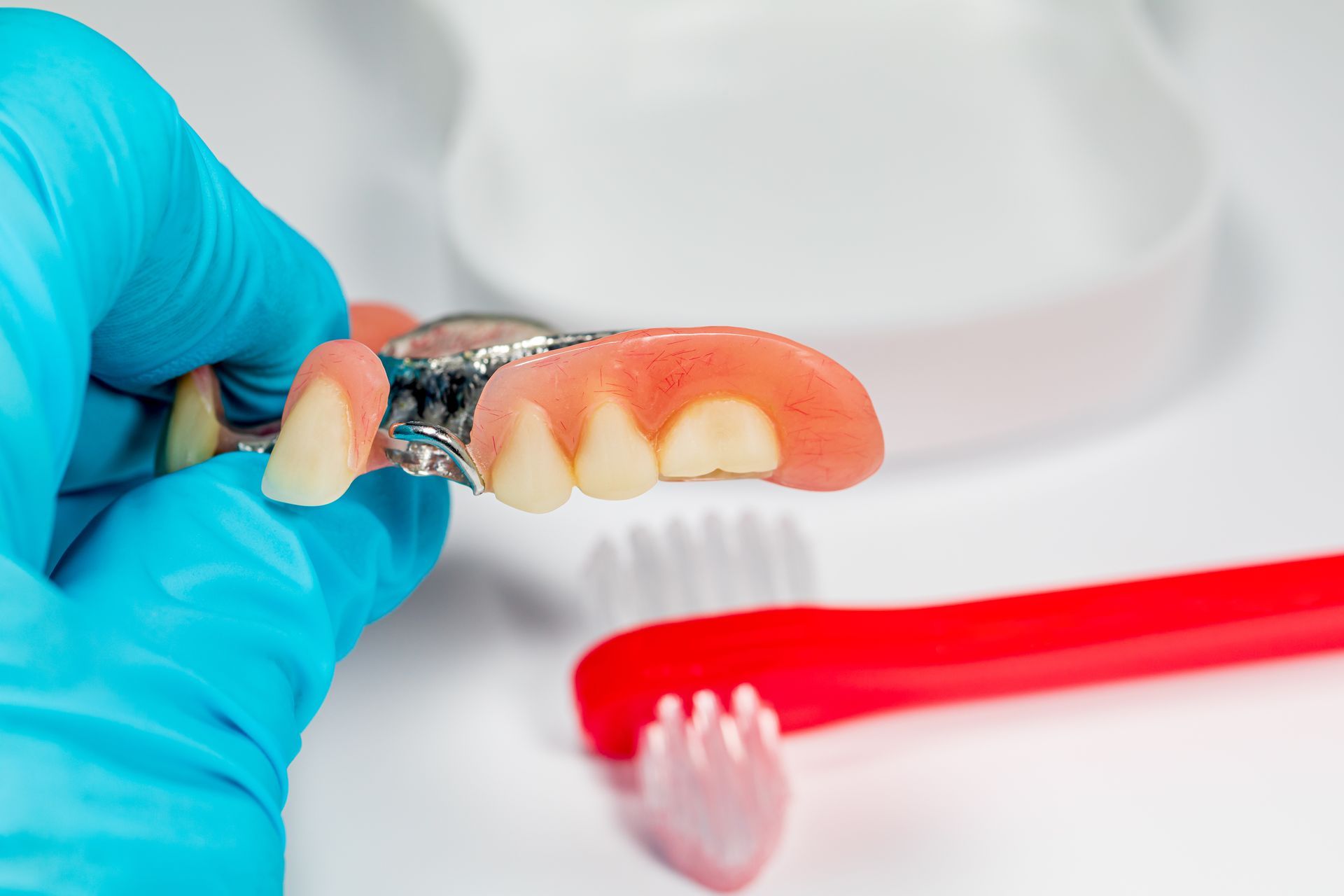 Gloved hand holding a partial denture beside a red toothbrush on a white surface