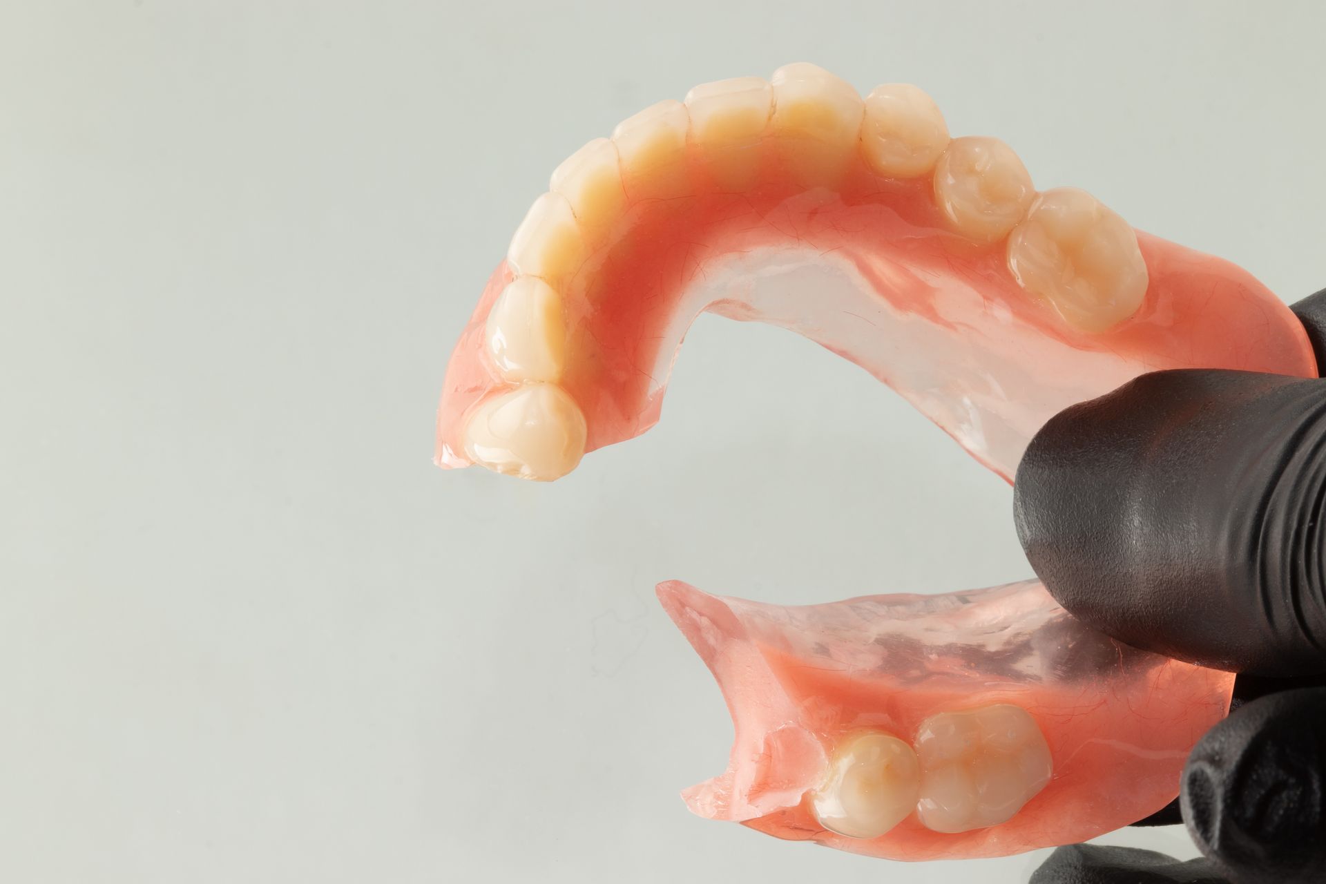 Gloved hand holding upper and lower pink dentures with white teeth on a light background