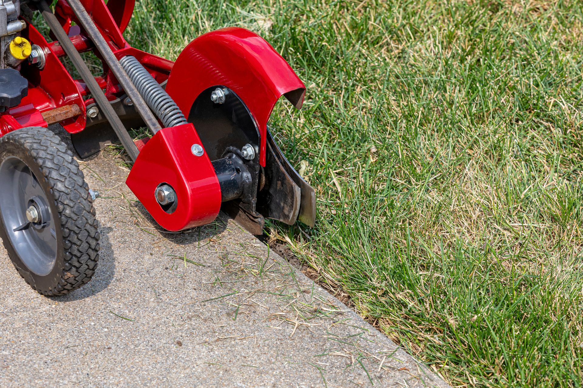 A red lawn mower is cutting the grass on the sidewalk.
