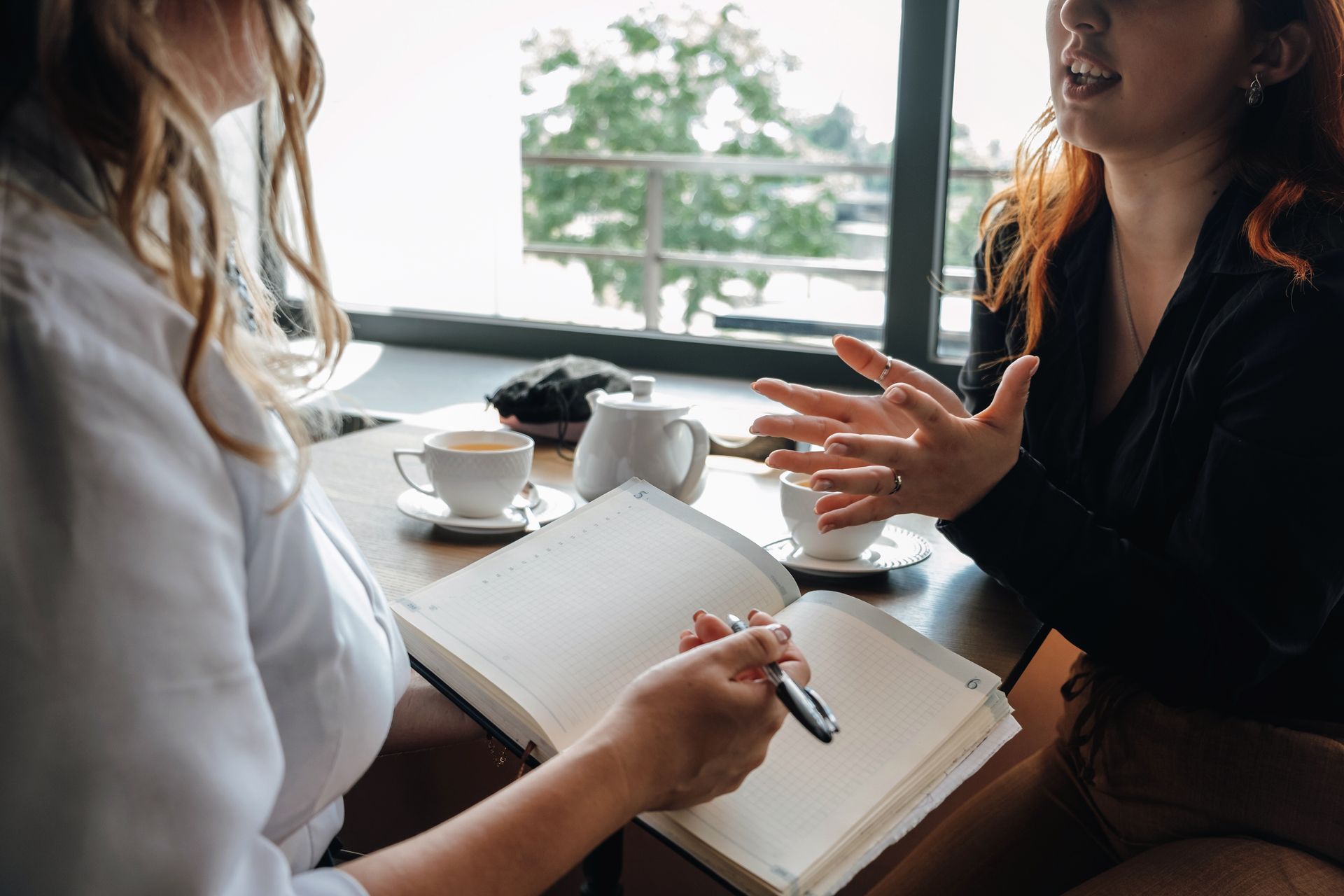 Two people talking at a café table, one writing in a notebook beside a coffee cup and teapot.