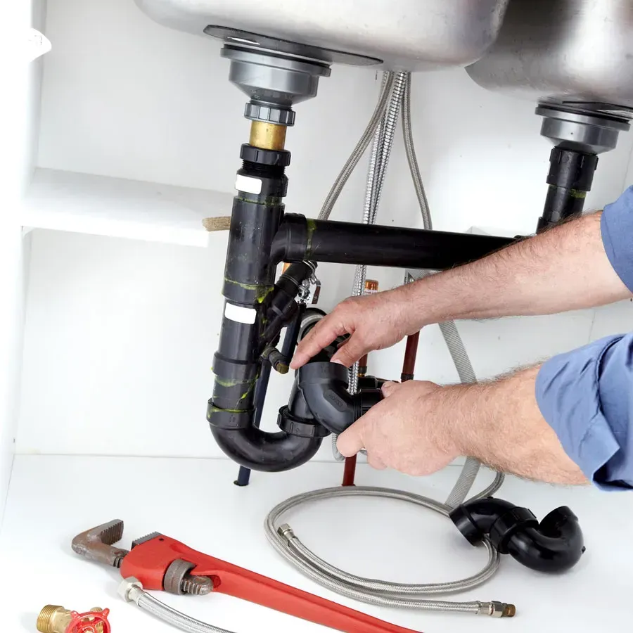 Plumber working under a kitchen sink, using a wrench. Black pipes, silver sink basins, and metal fixtures visible.